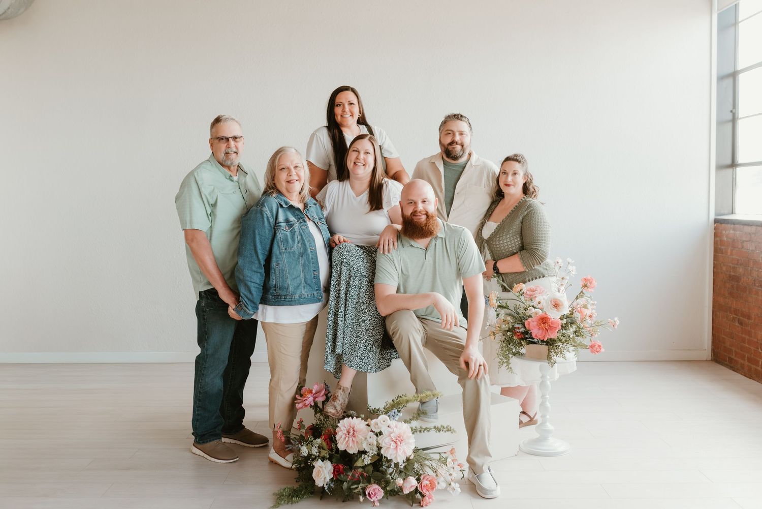Group of seven smiling adults pose together in bright white studio with pink floral arrangement.
