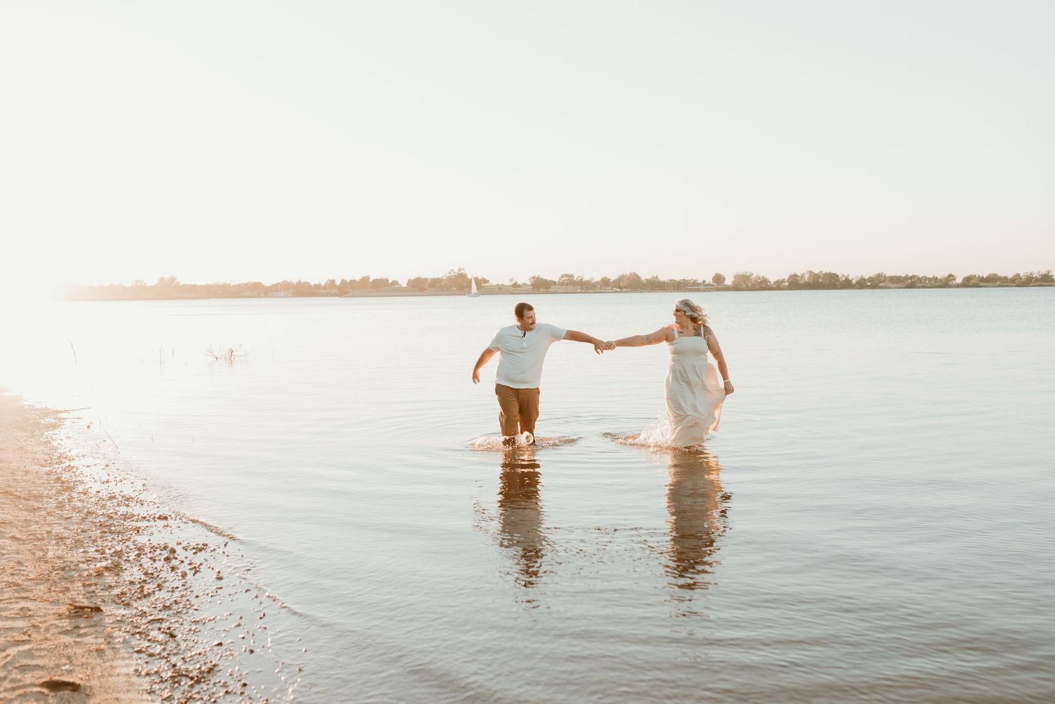 Joyful couple holding hands while walking through calm shallow water along a sunlit beach.