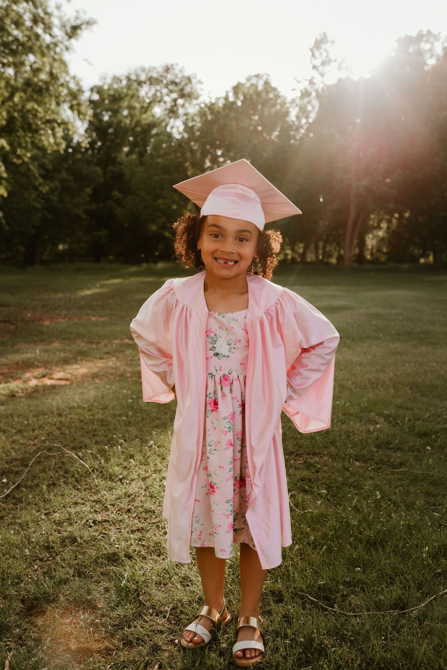Young girl proudly wearing pink graduation cap and gown standing outdoors in golden sunlight.
