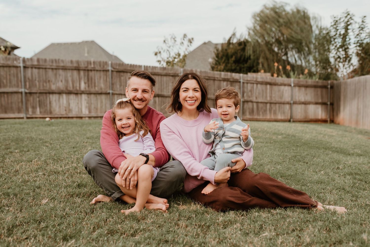 Happy family of four sitting on green grass in backyard, parents smiling with two young children.