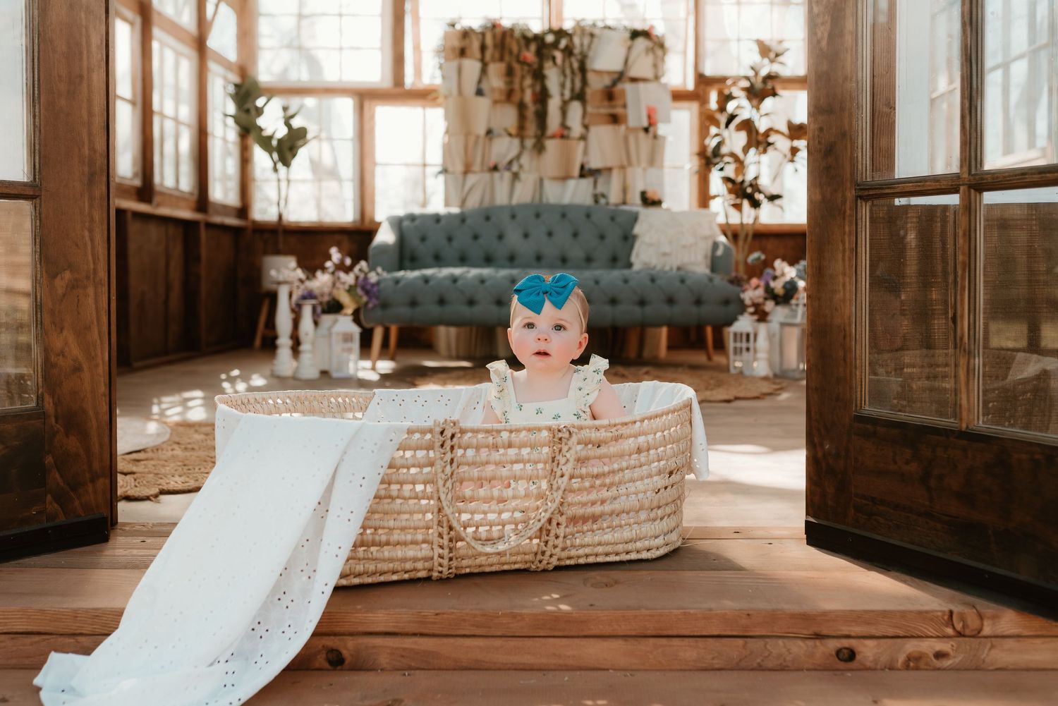 A baby in a wicker Moses basket sits on wooden floors inside a warmly decorated rustic studio.