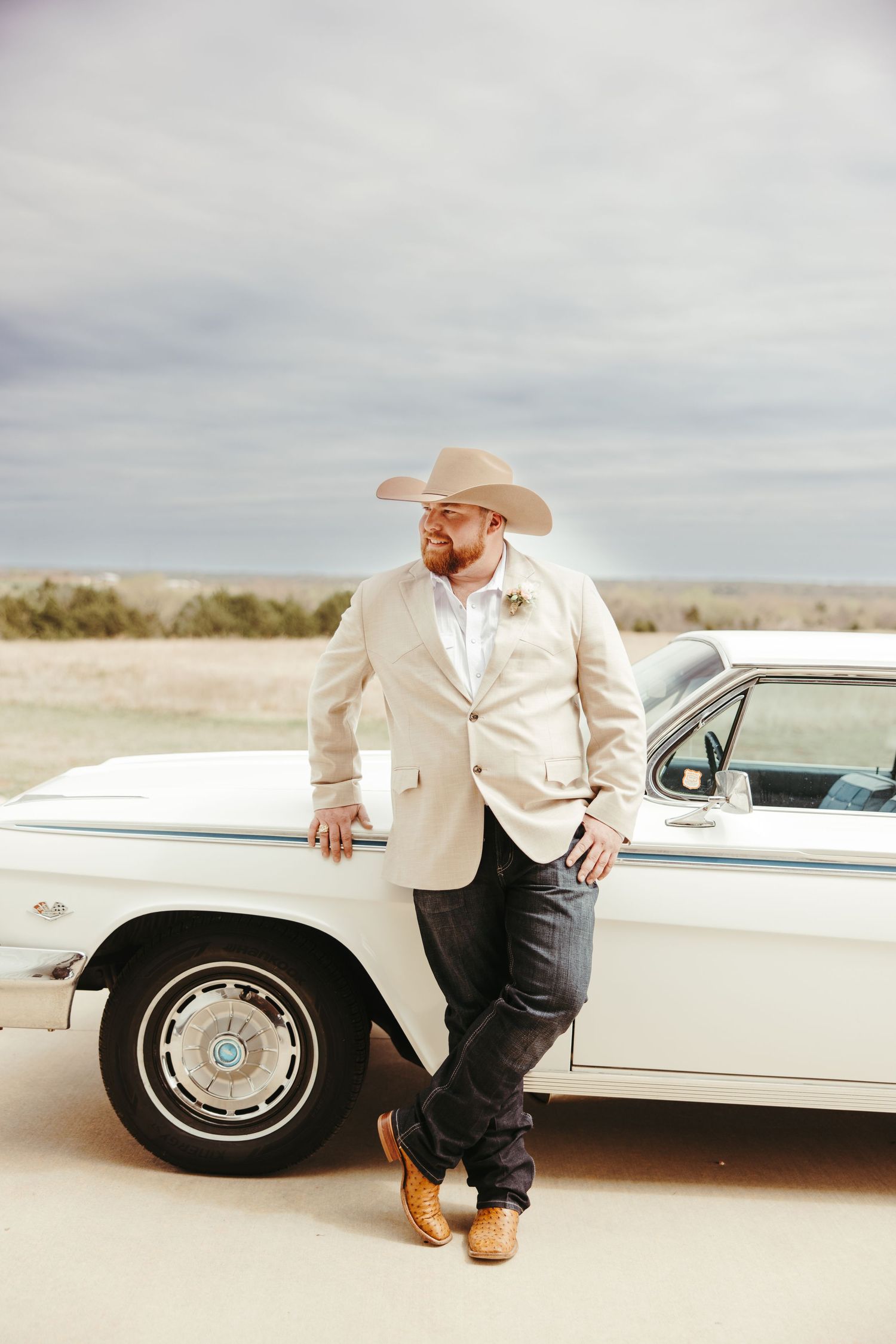 Groom in tan blazer and cowboy hat leans against a vintage white classic car under a cloudy sky.