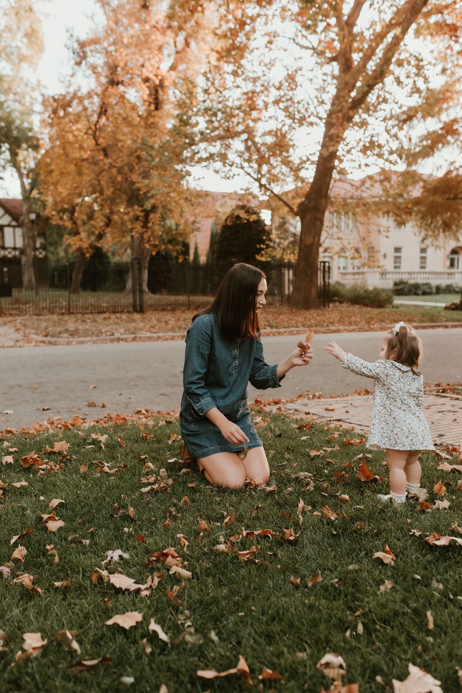 Mother in denim dress kneels on grass as toddler girl in floral dress hands her a fallen autumn leaf.