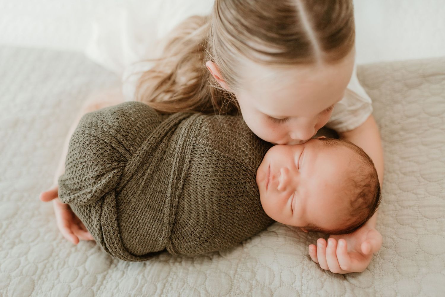 Young sibling gently kissing a sleeping newborn swaddled in an olive green knit wrap on a white blanket.