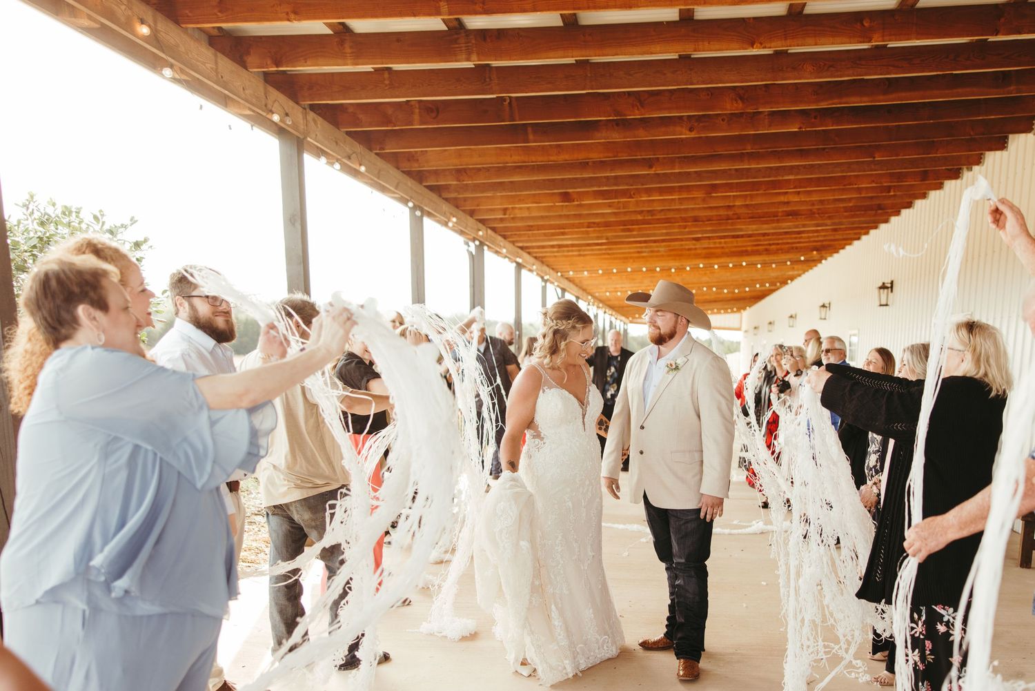 Bride and groom walk through a ribbon send-off under a covered outdoor venue surrounded by wedding guests.