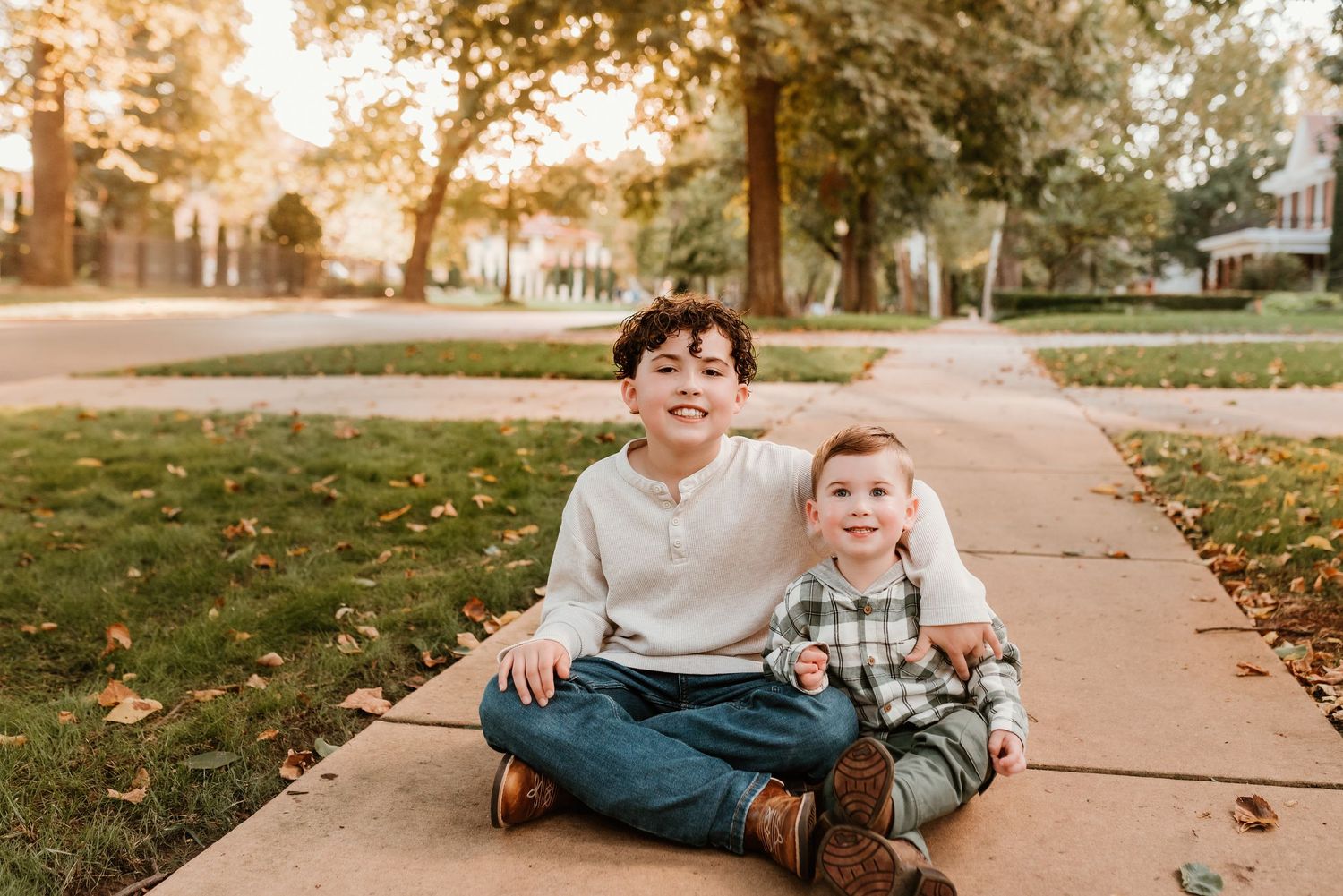 Two young brothers sitting together on a sunny sidewalk smiling during a fall family photo session.