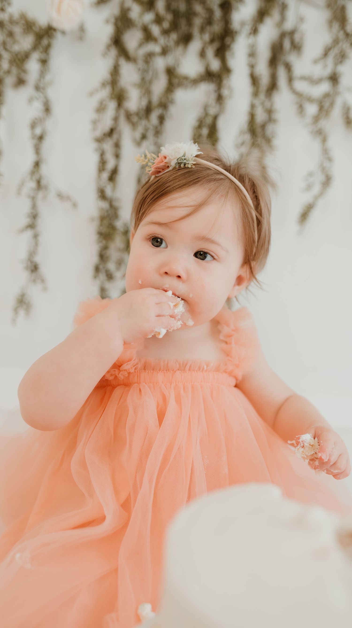 Adorable toddler in peach tulle dress with floral headband eating cake during first birthday smash session.