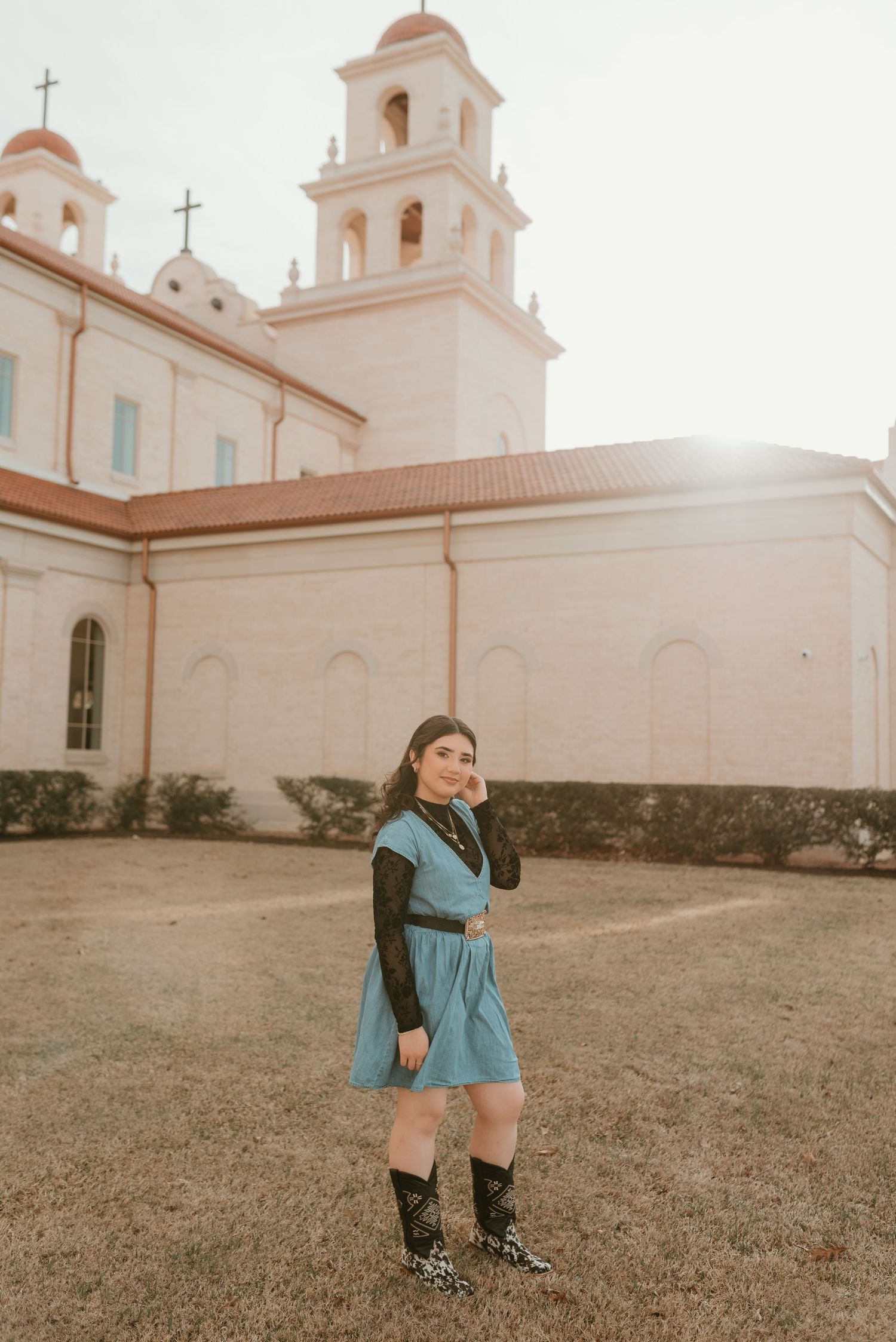 Young woman in blue dress and cowboy boots posing in front of a sunlit white mission church.