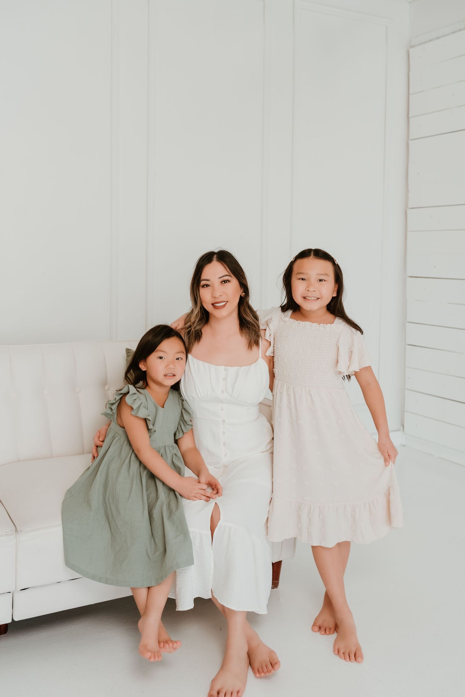 Asian mother in white dress posing with two young daughters in bright white studio setting.