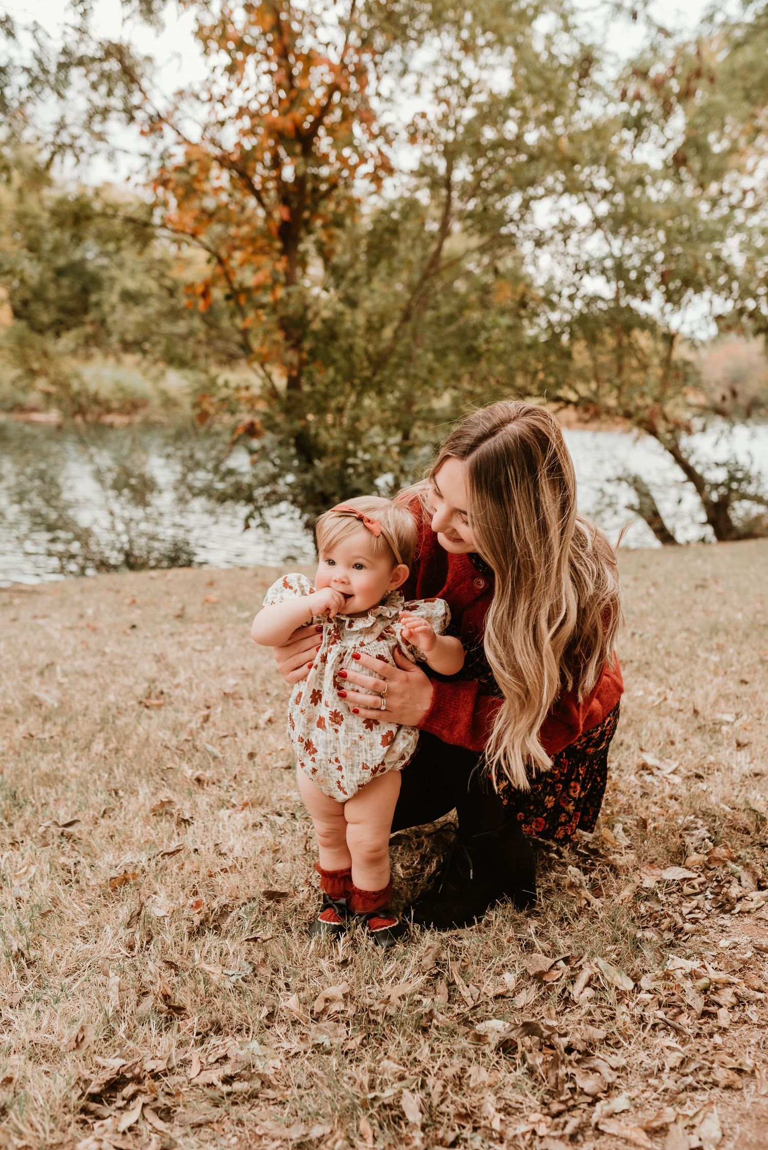 Mother crouches beside her baby girl near a serene autumn lake surrounded by fallen leaves.