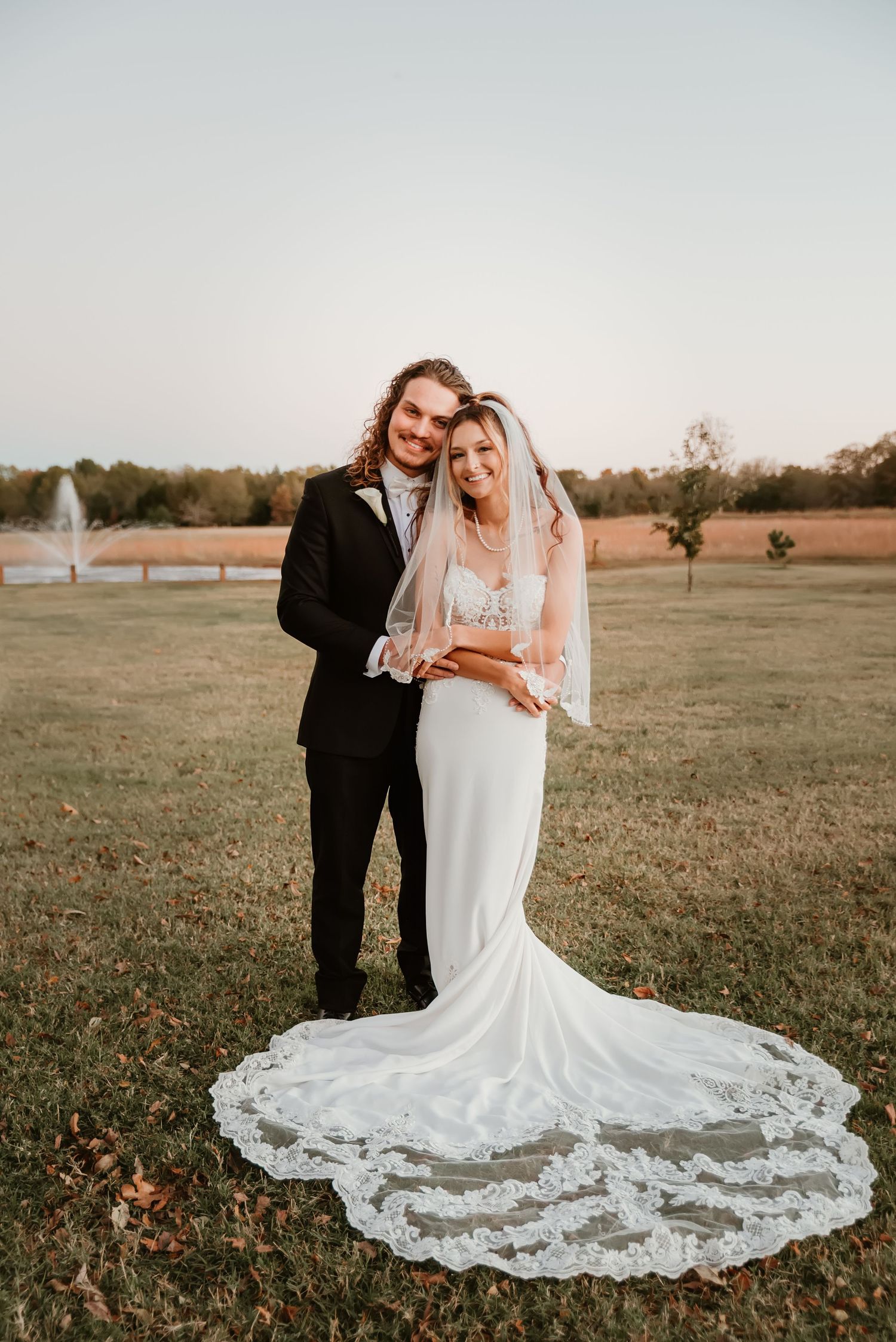 Bride in lace mermaid gown with cathedral veil and groom in black tuxedo pose together at golden hour outdoors.
