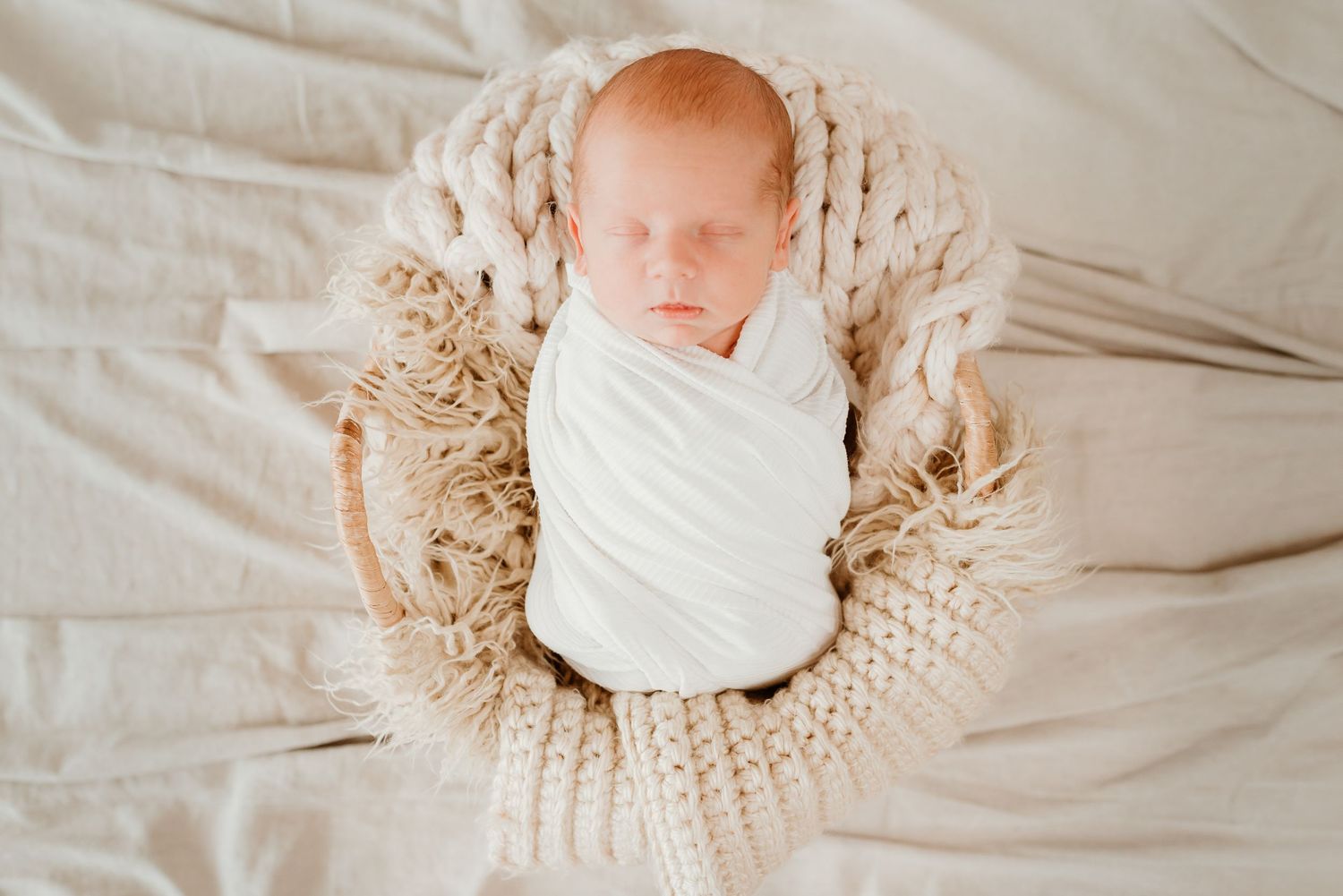Sleeping newborn baby swaddled in white wrap nestled in a chunky knit basket bowl.