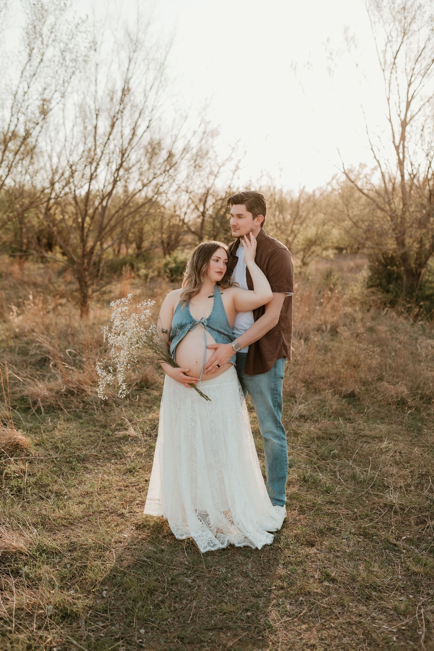 A couple embraces in a golden field at sunset, the woman in a flowing white skirt cradling her bump.