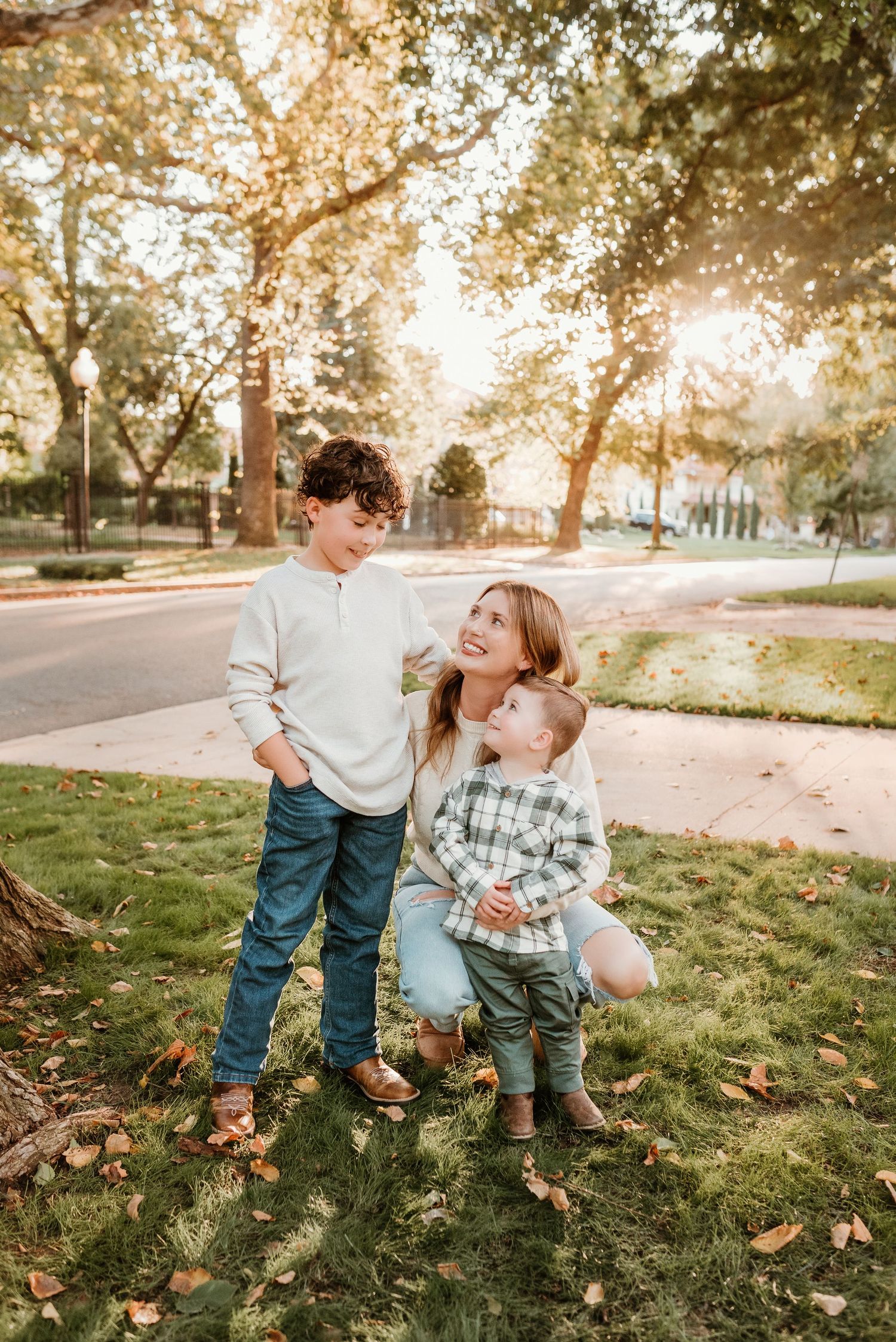 Mother kneels on grass smiling at two young sons during golden hour outdoor family portraits.