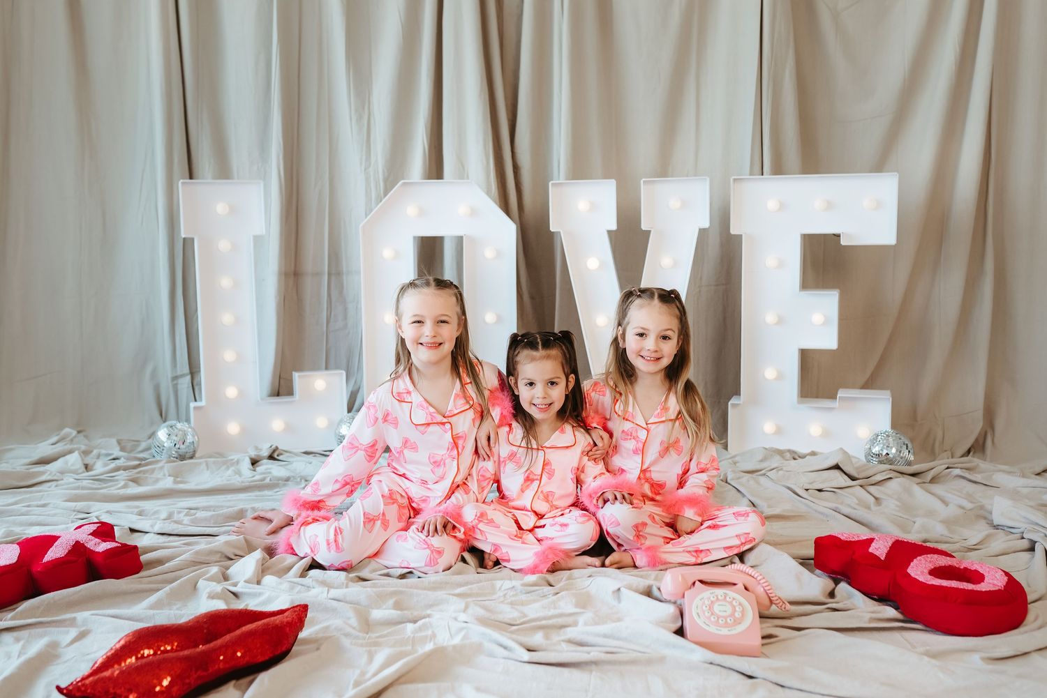 Three girls in pink pajamas sit together in front of illuminated LOVE letters for Valentine's Day.