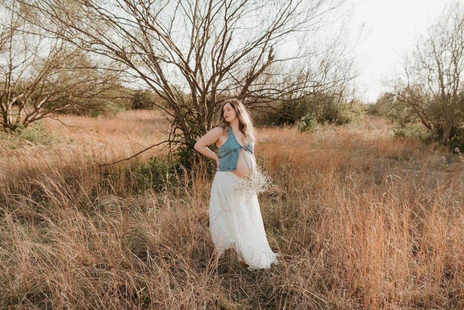 A pregnant woman in a denim top and flowing white skirt poses gracefully in a golden outdoor field.