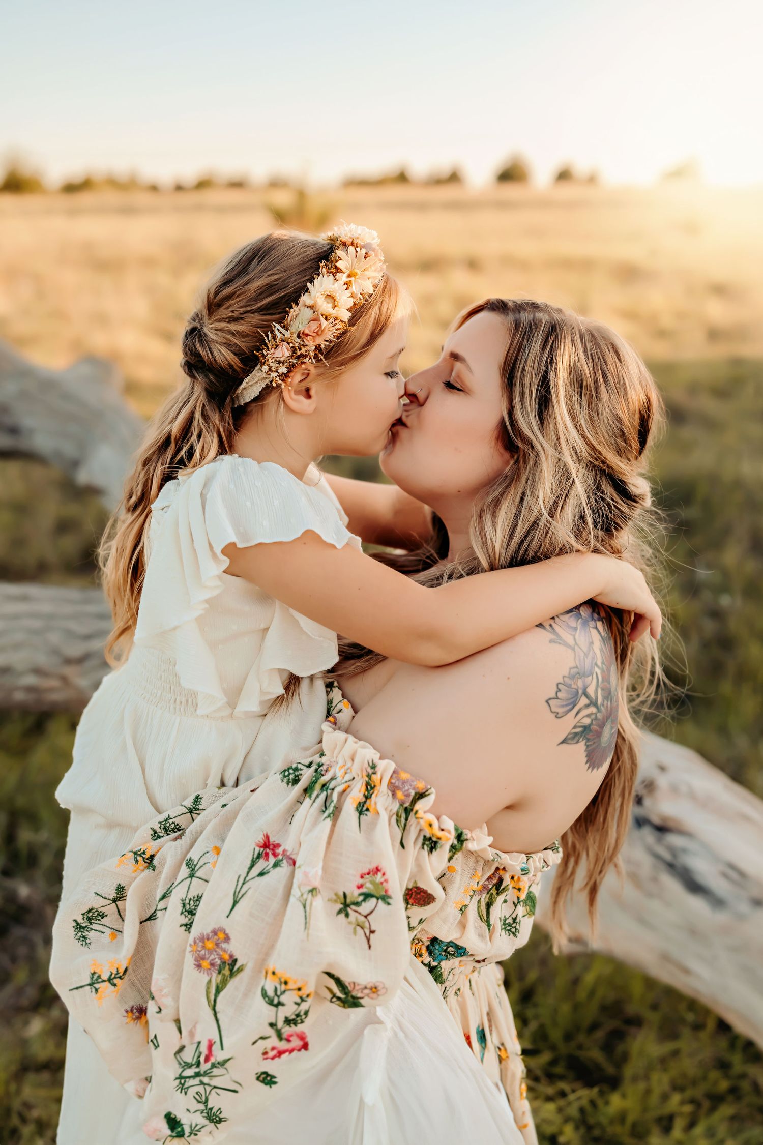 Pregnant tattooed mother kissing young daughter wearing floral crown in golden sunset field.