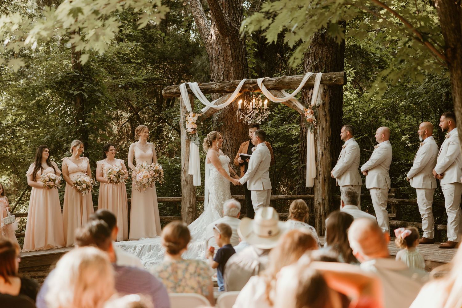 Outdoor woodland wedding ceremony with couple at floral arch, bridesmaids in blush and groomsmen in tan.