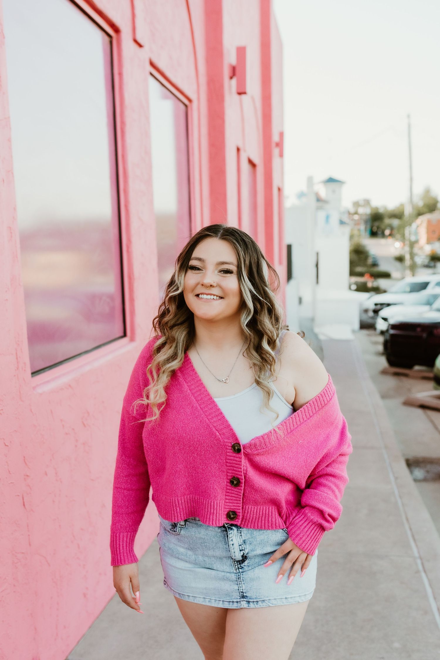 Young woman in hot pink cardigan and denim skirt smiling beside pink building on sidewalk.