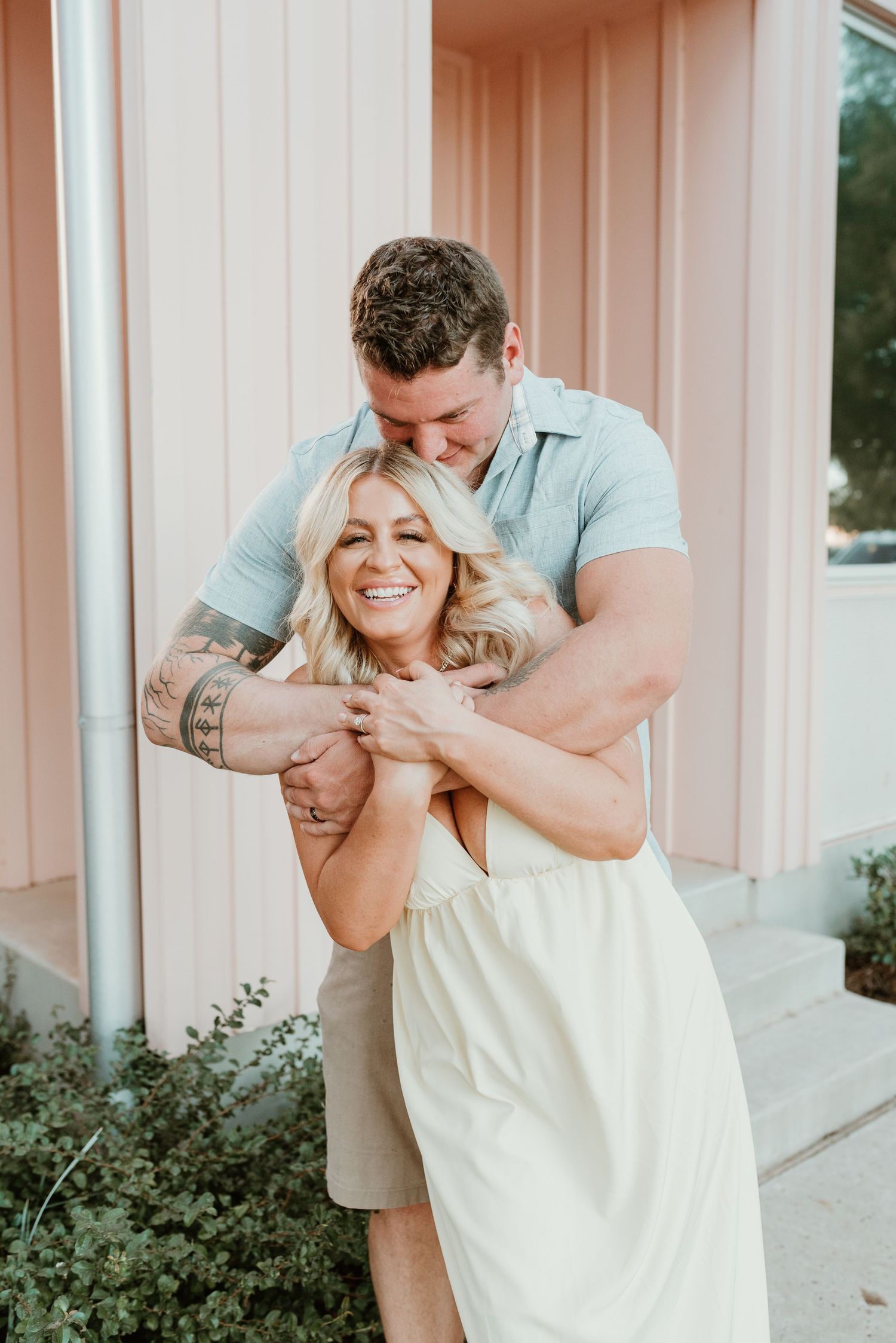 A tattooed man hugs a laughing blonde woman in a white dress against a pink building exterior.