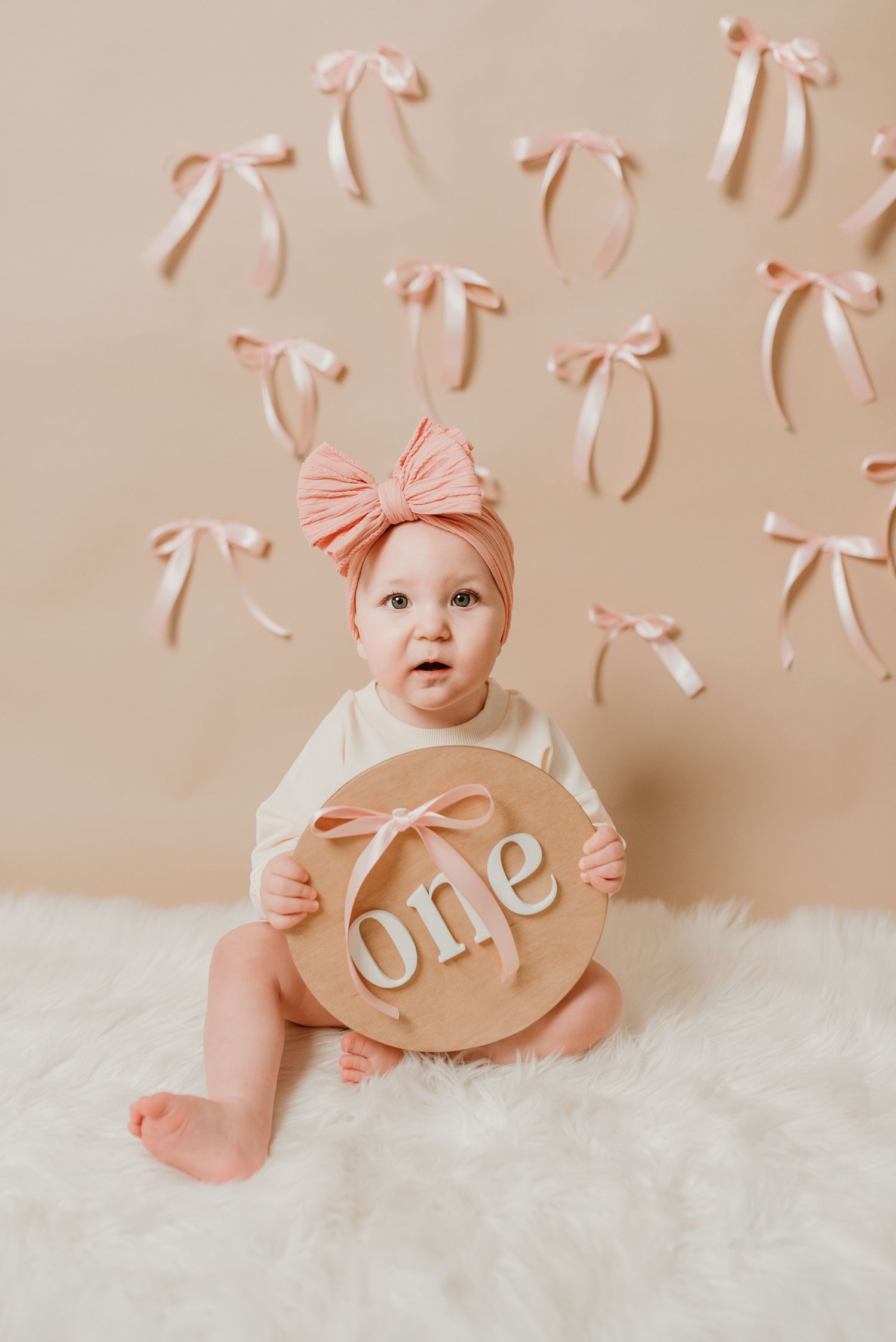 Cute baby girl in pink bow headband holding wooden one sign on fluffy white rug.