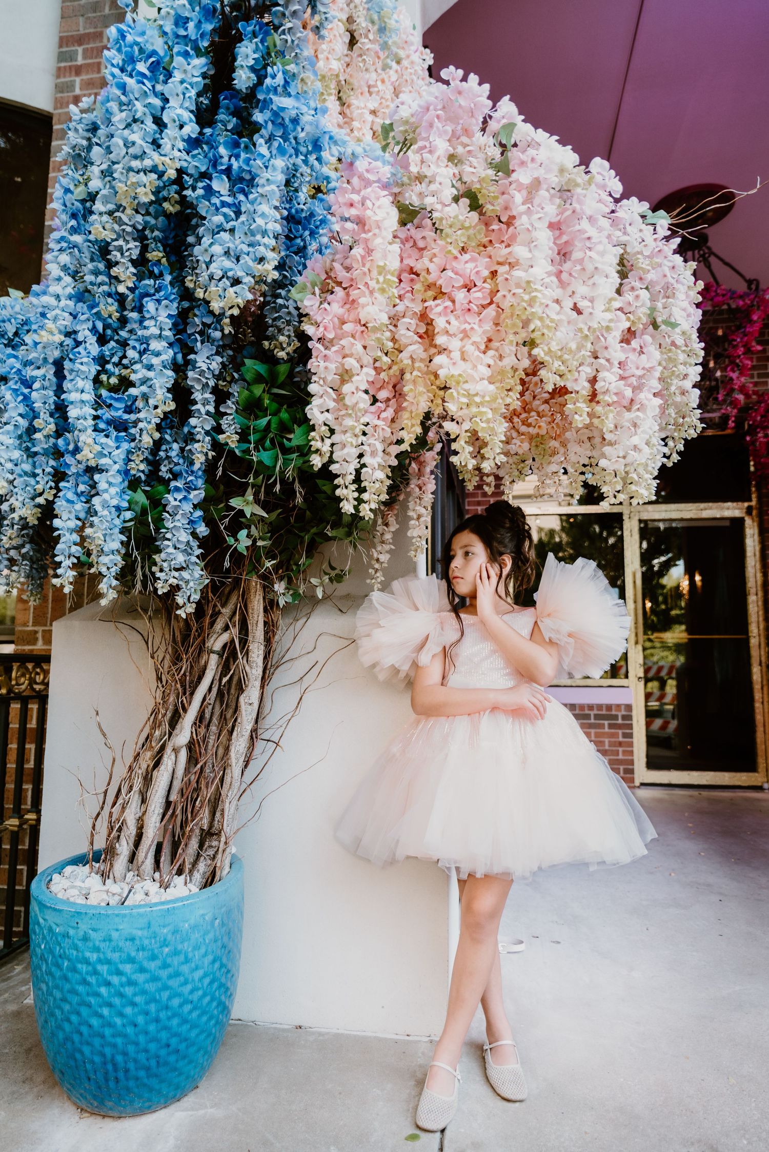 Young girl in pink tutu dress standing under colorful hanging wisteria floral display outdoors.