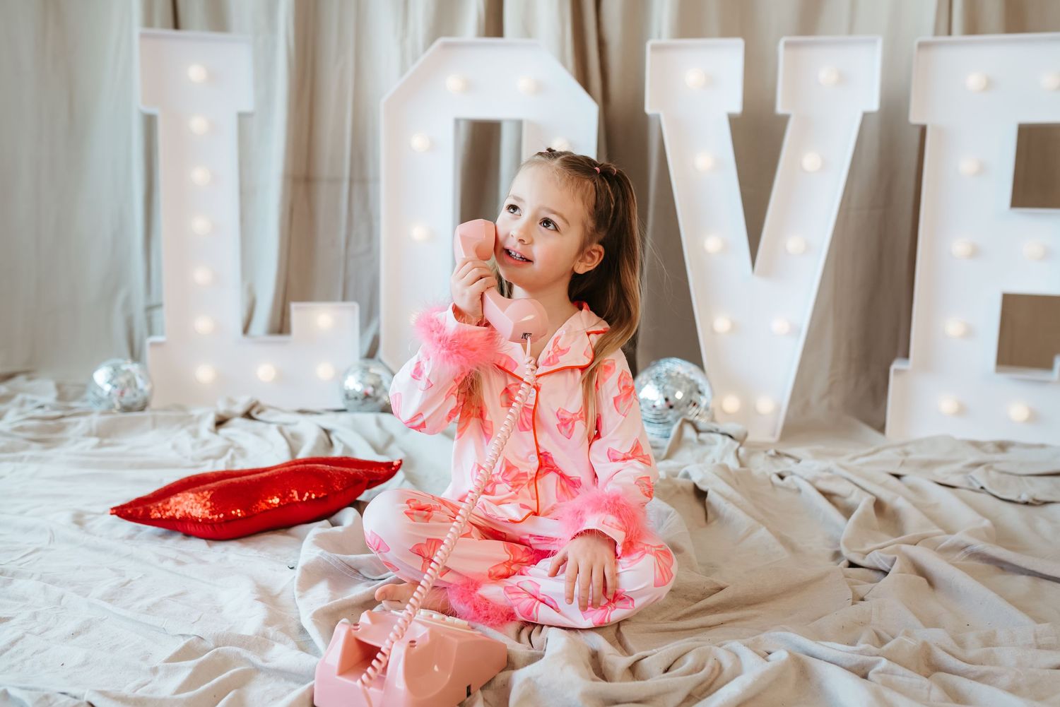 A young girl in pink pajamas sits playfully with a toy phone before glowing LOVE letters.