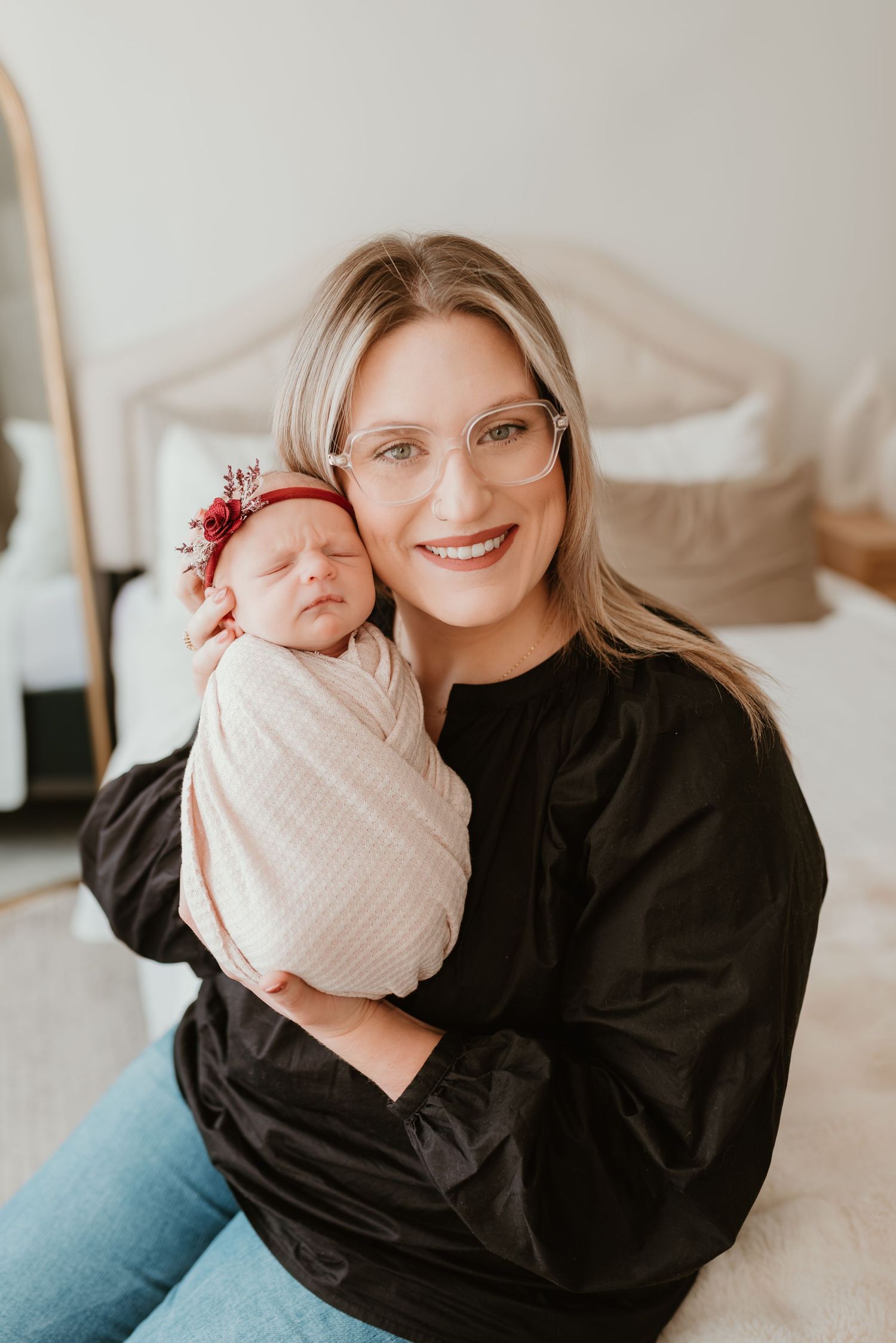 Smiling blonde mother with glasses holding sleeping newborn with red floral headband indoors.