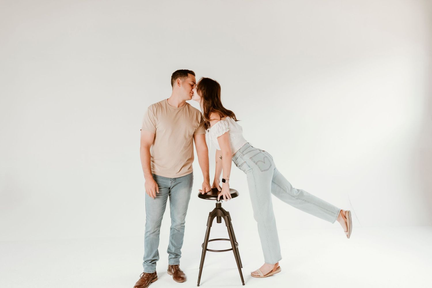 Couple sharing a tender kiss in a bright studio, woman playfully lifting her leg beside a wooden stool.