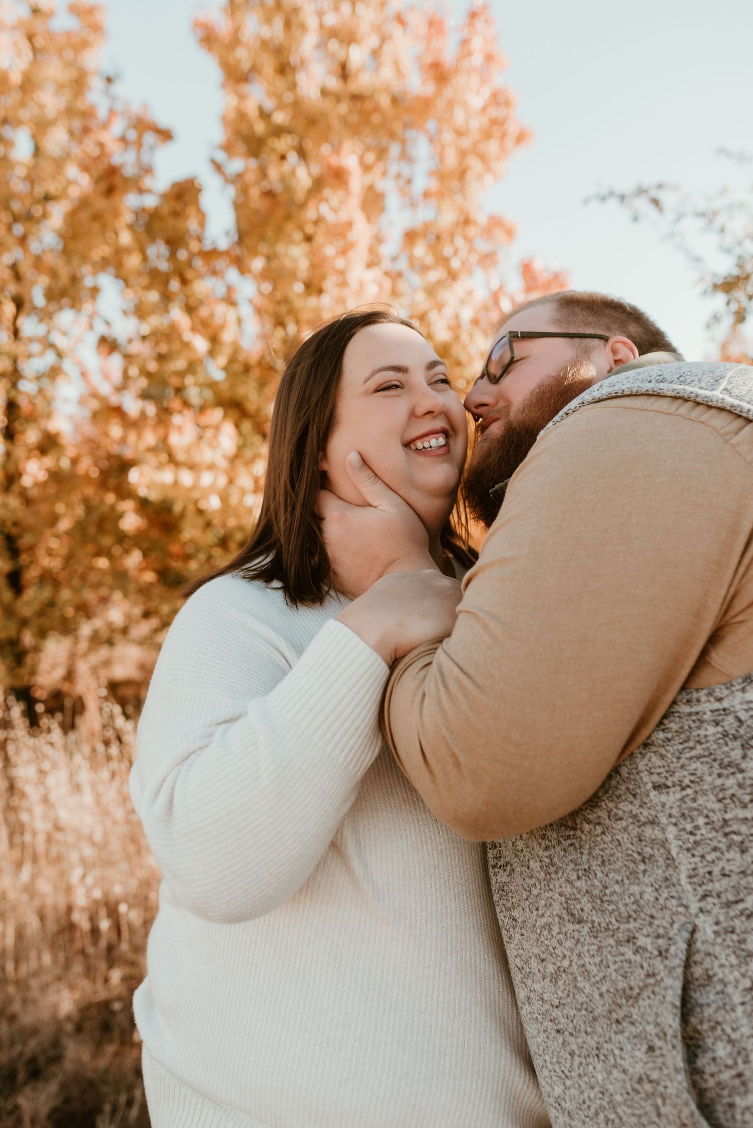 Happy couple sharing a tender moment outdoors among autumn foliage, woman in cream sweater laughing joyfully.