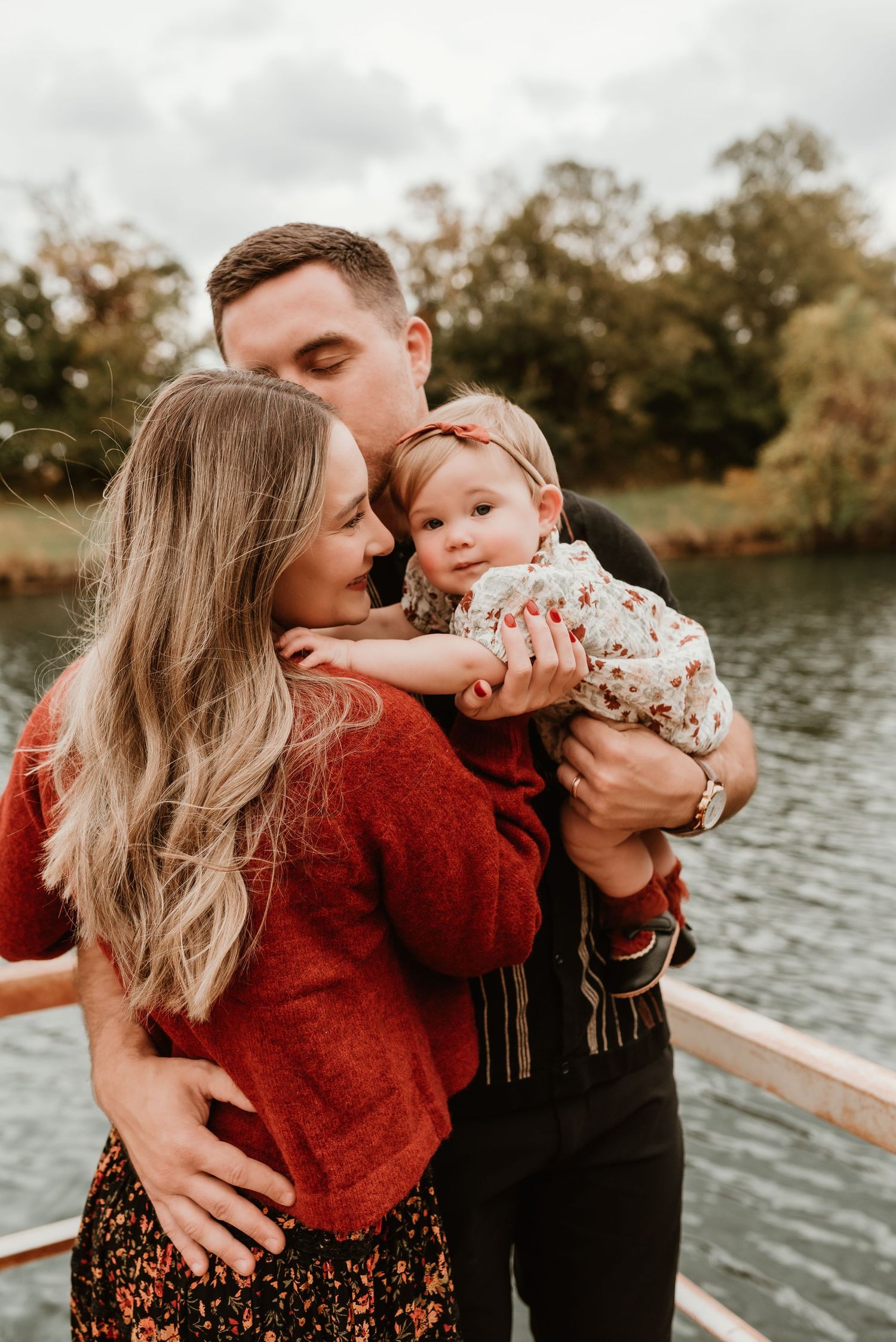 A couple shares a tender moment holding their baby girl near a scenic lakeside on a cloudy day.