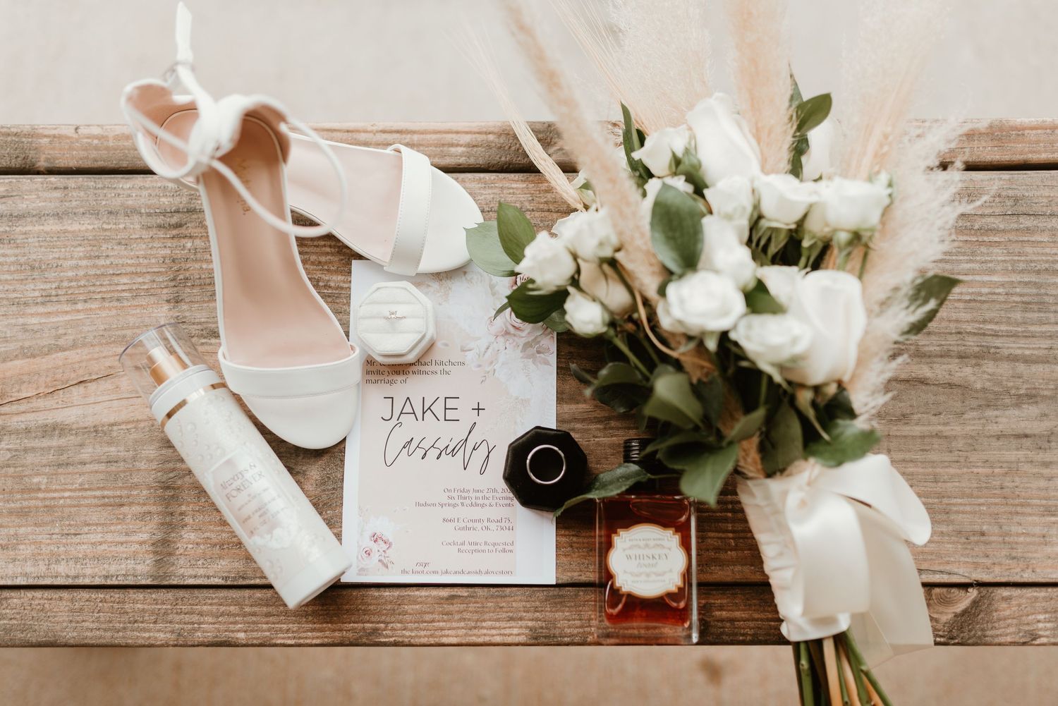 Flat lay of wedding details including white heels, roses, rings, perfume, and Jake and Cassidy invitation.