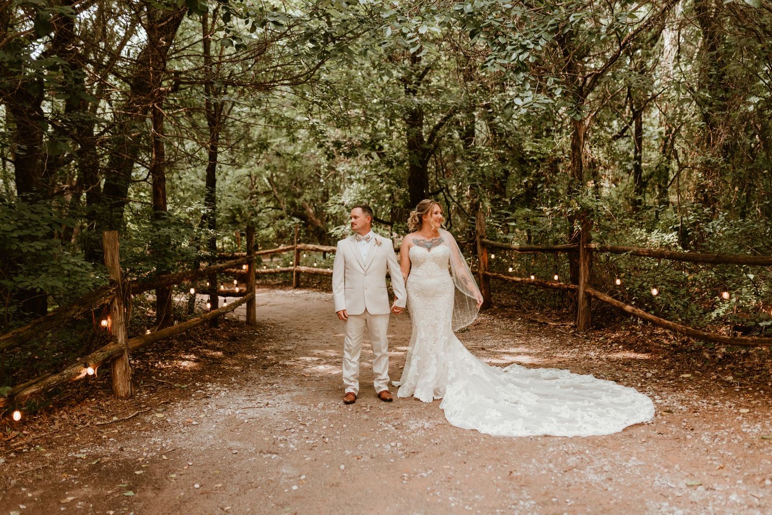 Bride and groom walking hand in hand on a wooded path lined with warm fairy lights.