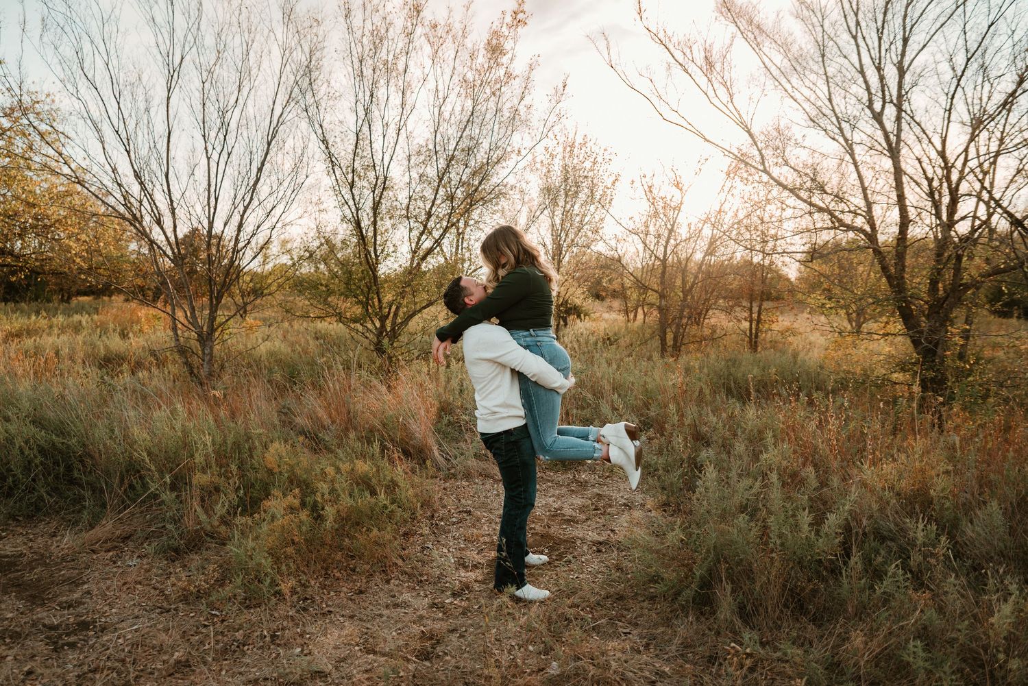 Man lifting woman in jeans and white sneakers in a joyful embrace in an autumn outdoor setting.