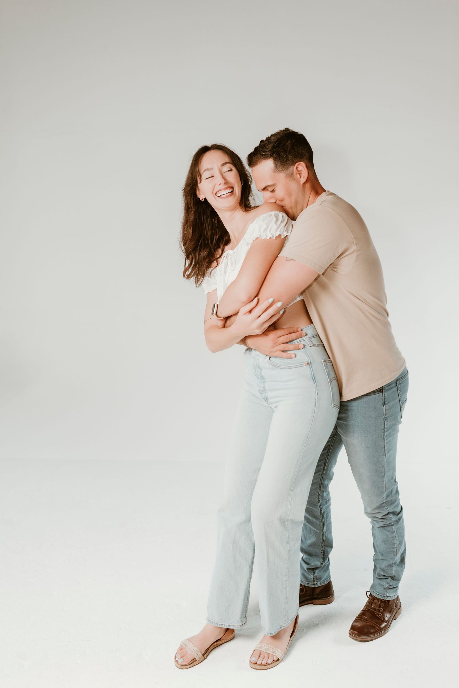 A laughing woman in white top and jeans is embraced from behind by a man in a beige shirt.
