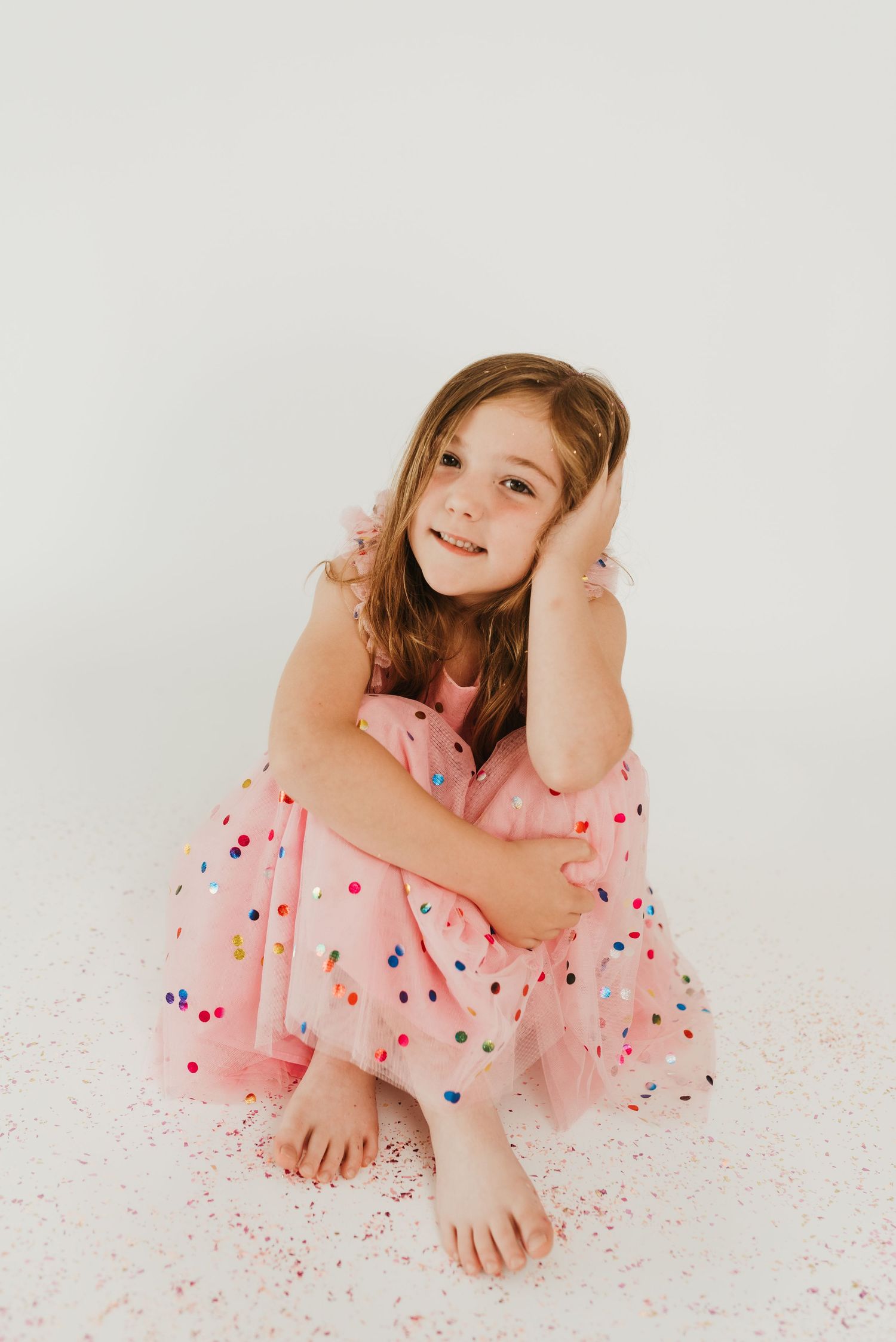 A young girl in a pink polka dot dress sits barefoot on the floor smiling warmly.
