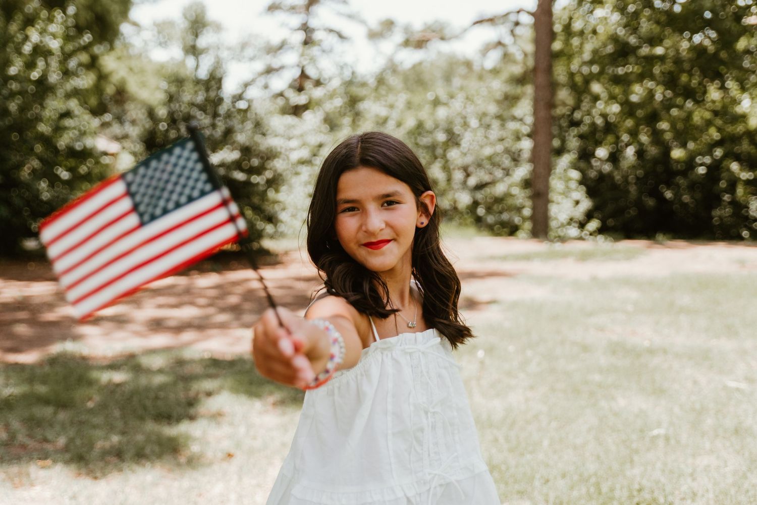 Young girl in white dress holds an American flag outdoors in a green park setting.