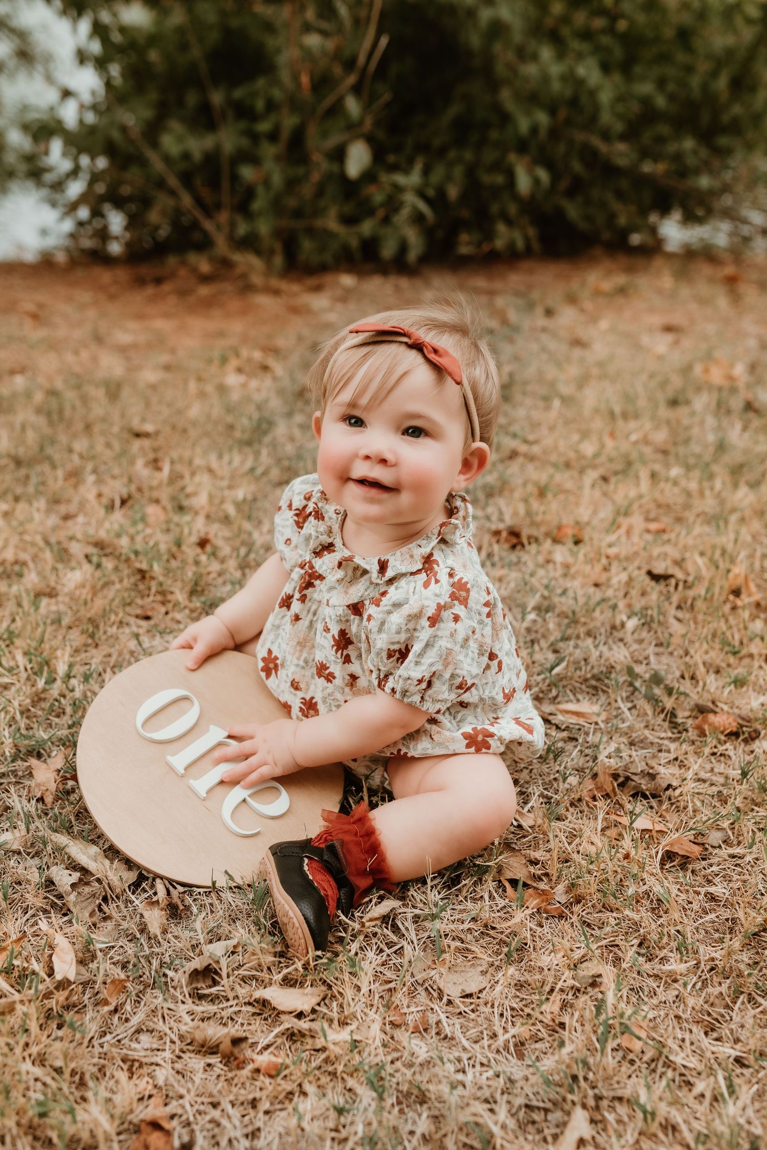 Smiling baby girl in floral romper sits on grass holding a wooden 'one' milestone sign.