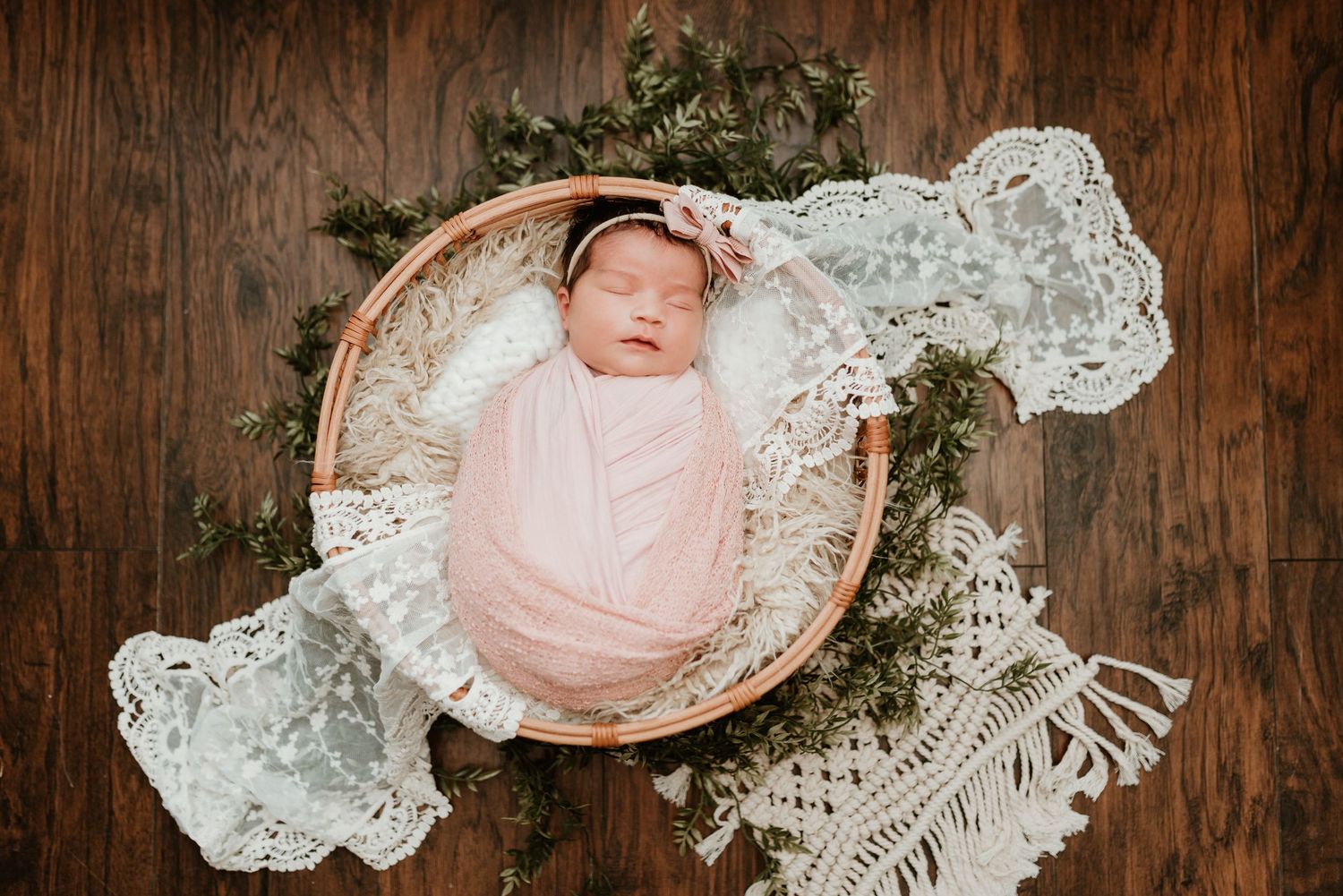 Sleeping newborn baby wrapped in pink swaddle inside a wooden bowl with lace and greenery.