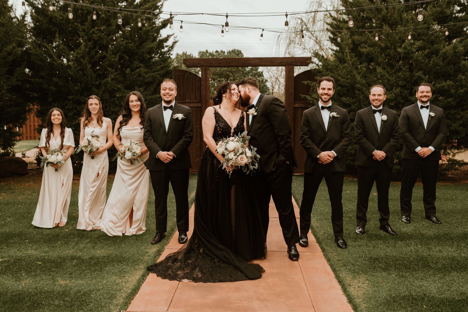 Wedding party poses with kissing bride and groom on outdoor aisle with string lights and wooden arch.