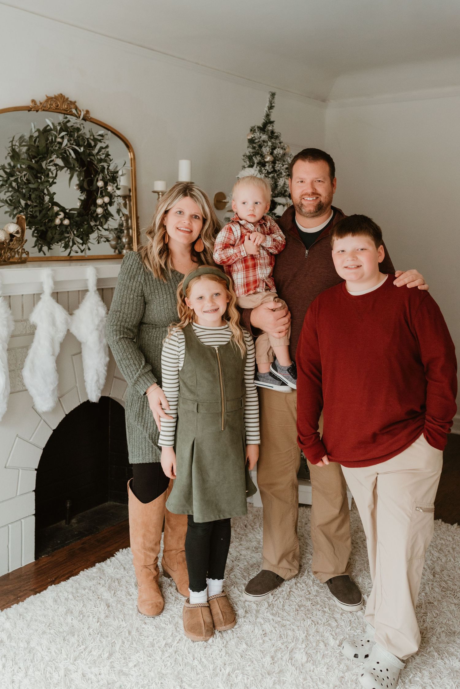 A family of five poses together in front of a fireplace and Christmas tree in holiday outfits.