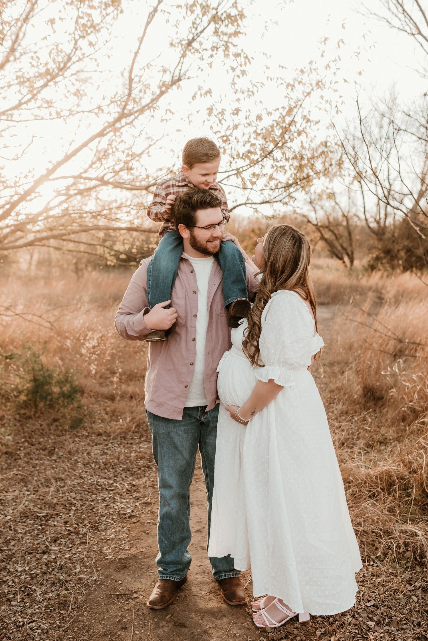Family maternity photo with pregnant woman in white dress, man with child on shoulders outdoors.