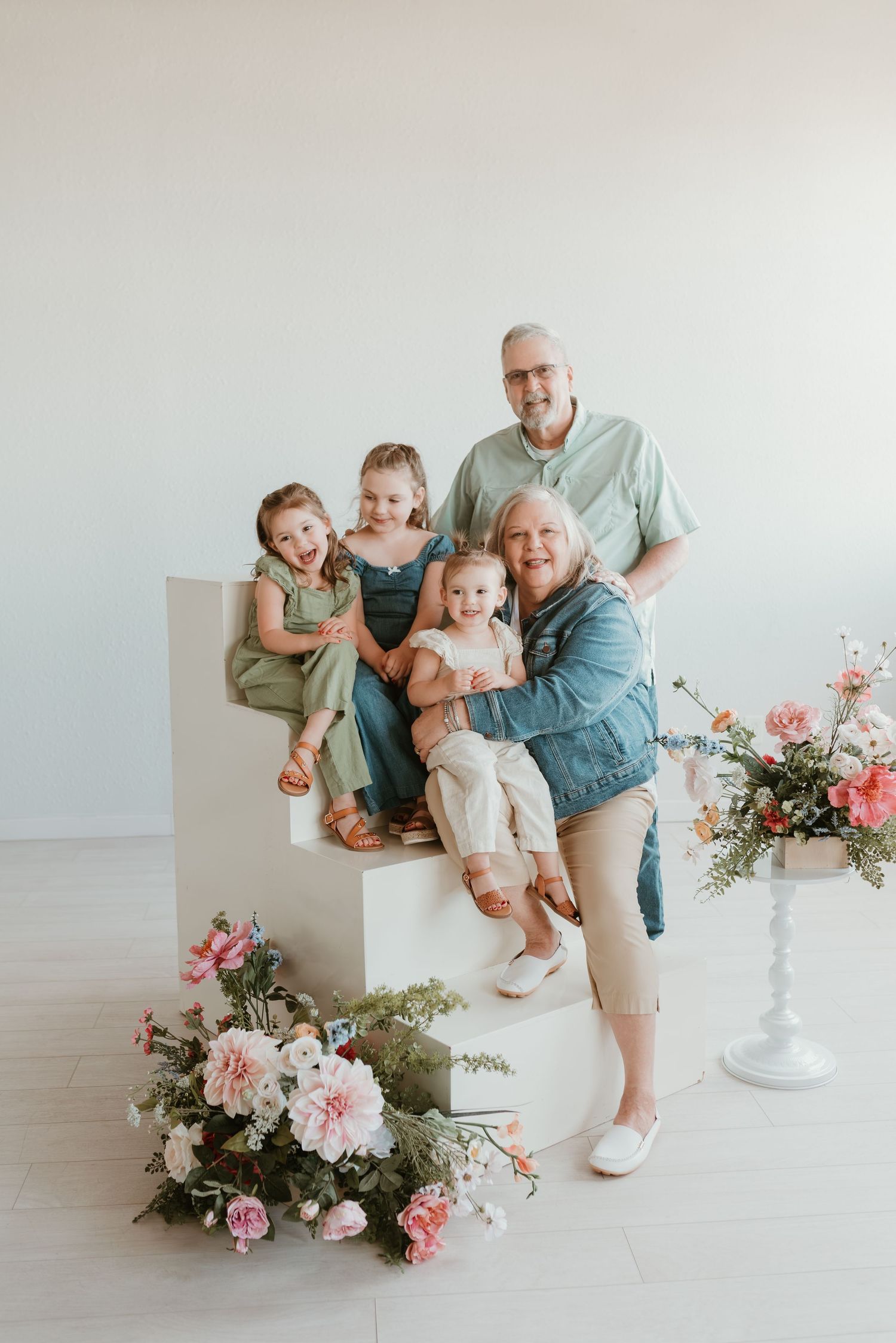 Grandparents pose joyfully with three young granddaughters surrounded by pink flowers in white studio.