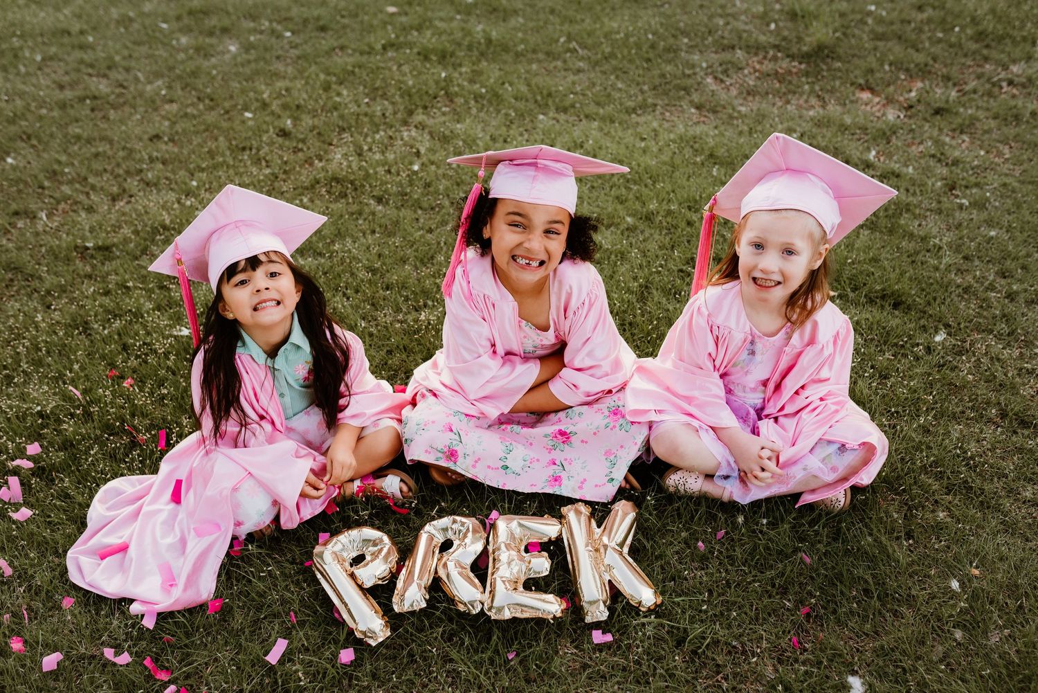 Three girls in pink cap and gown sitting on grass with gold PREK balloon letters for preschool graduation.