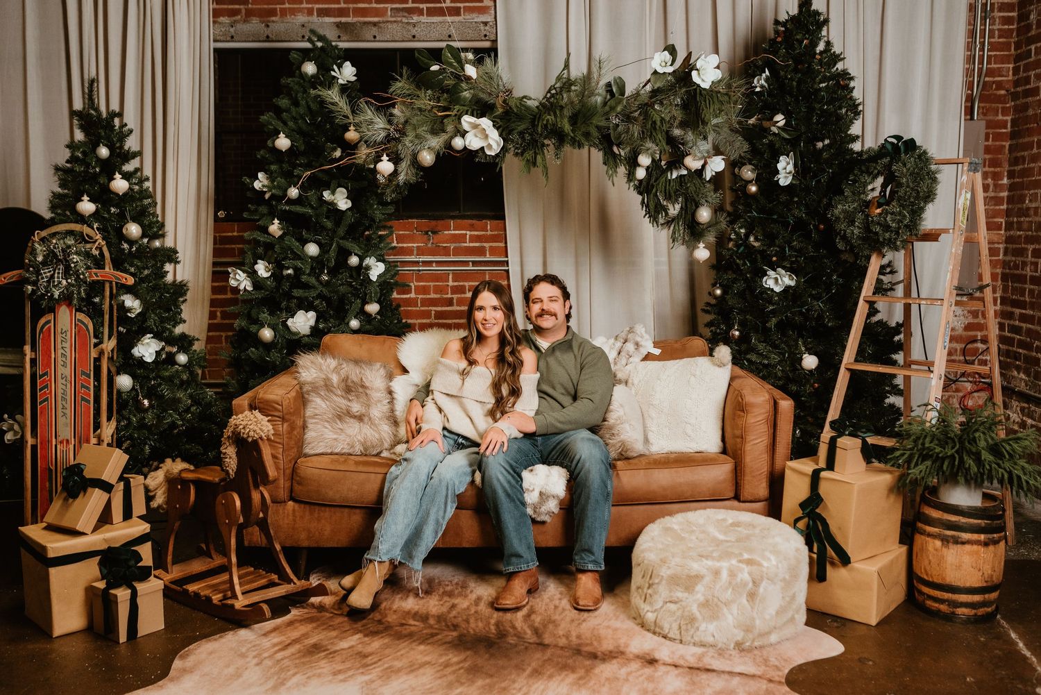 A smiling couple sits on a leather sofa surrounded by Christmas trees and holiday decorations indoors.