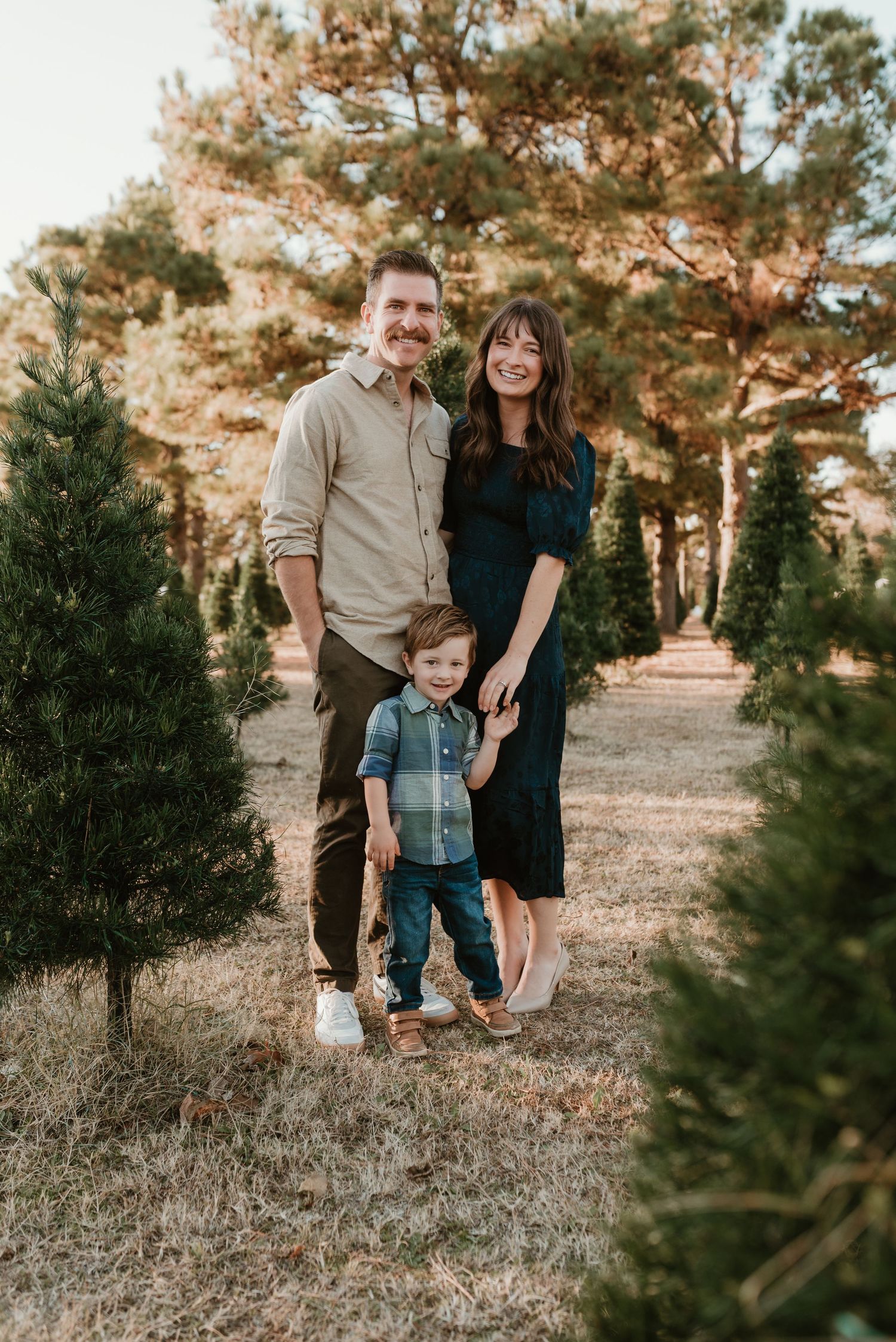 Smiling family of three stands among Christmas trees at a festive tree farm.