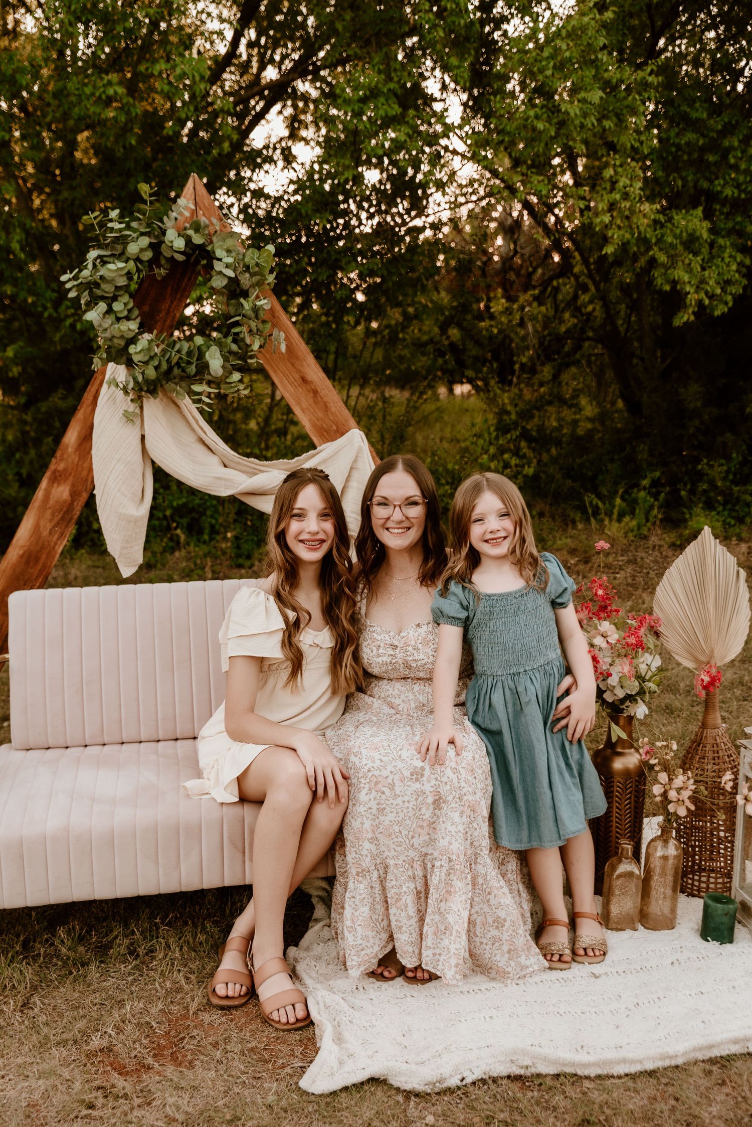 Woman with two daughters poses on pink couch under boho arch with greenery wreath backdrop.