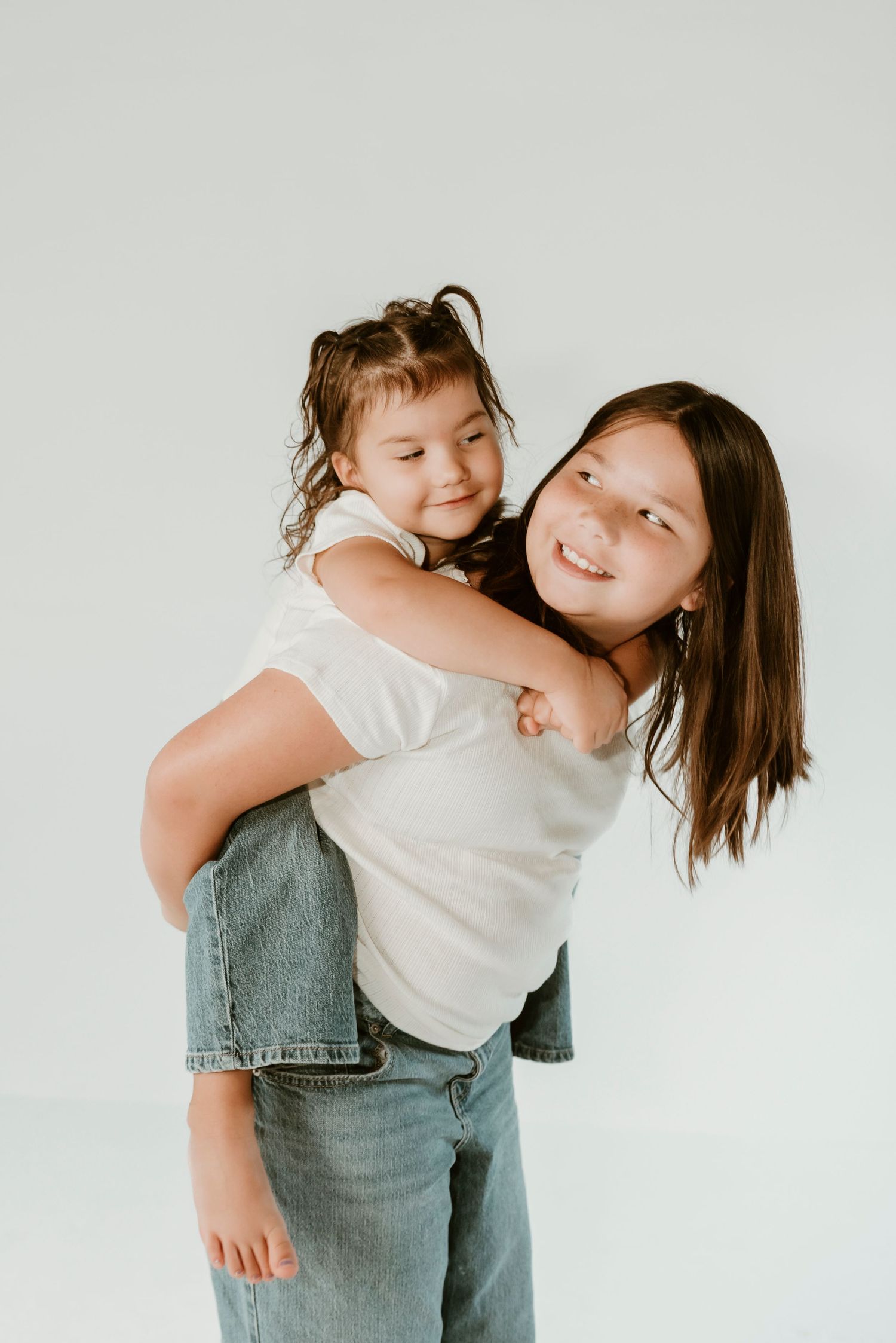 A smiling older girl gives a laughing younger girl a piggyback ride against a white background.