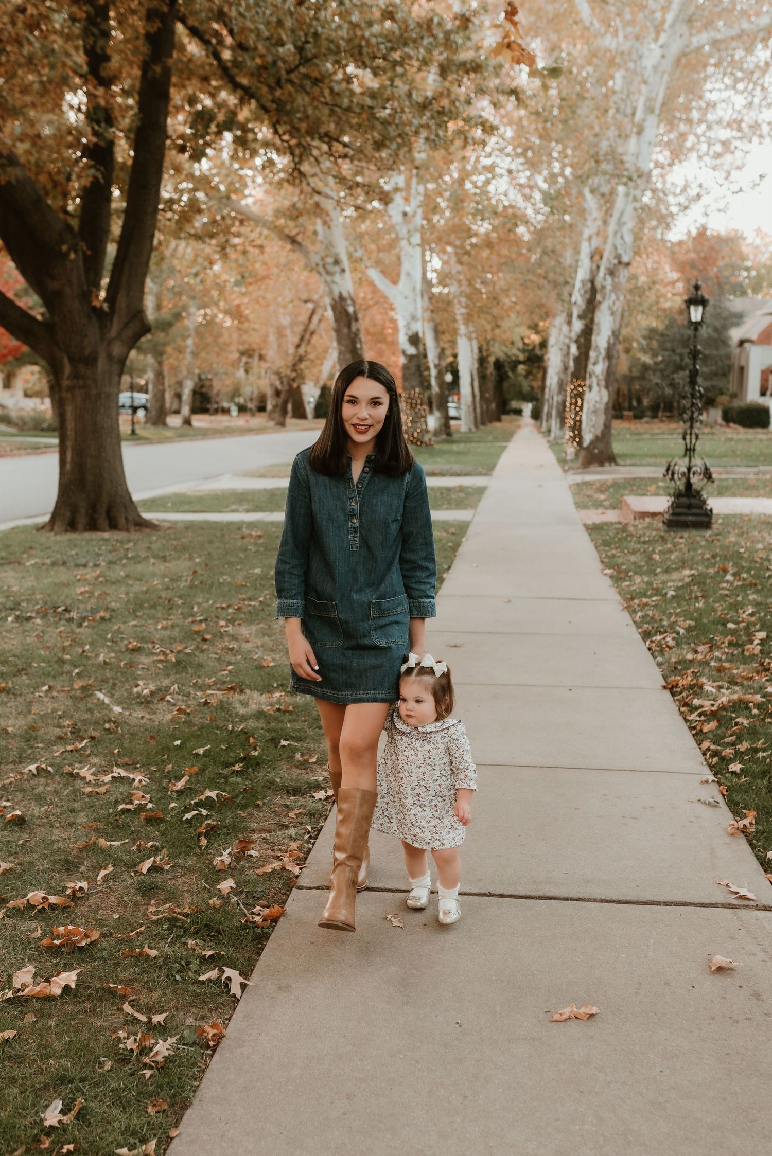 Mother in denim dress walks with toddler daughter in floral outfit along a tree-lined sidewalk in autumn.