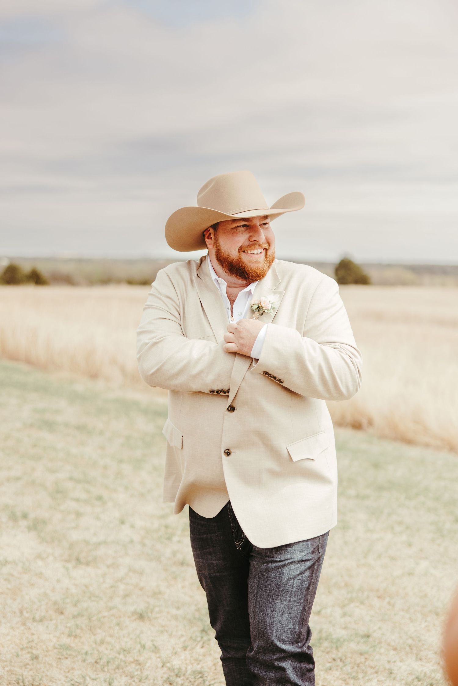 Smiling groom in cream blazer and cowboy hat adjusts boutonniere while standing in a golden field.